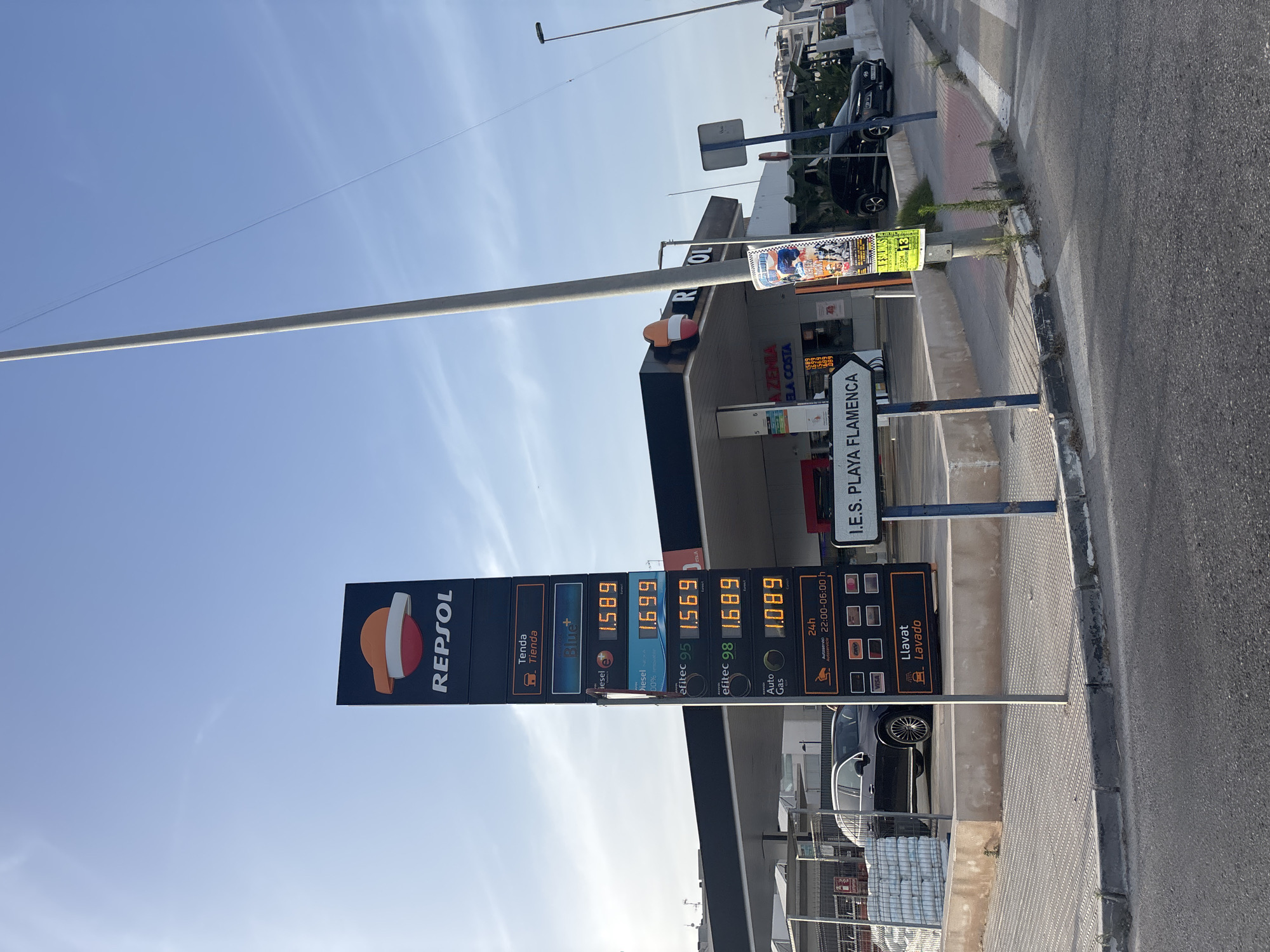 The image shows a Repsol gas station in Orihuela, Spain. The gas station features a prominent Repsol logo and a digital price board displaying fuel prices. The prices listed are for "Tenda" (Store), "Blue+", "diesel e", "diesel 00%", "efitec 95", "efitec 98", and "Auto Gas". The "Auto Gas" price is 1.089, and the "efitec 95" price is 1.569. The station appears to be open, with cars present, including a dark-colored SUV. A sign indicating "I.E.S. PLAYA FLAMENCA" is visible, suggesting the gas station is located near this educational institution. The environment is outdoors, with a clear blue sky and sunlight casting shadows. The scene is captured from a street-level perspective, with a road and sidewalk in the foreground.