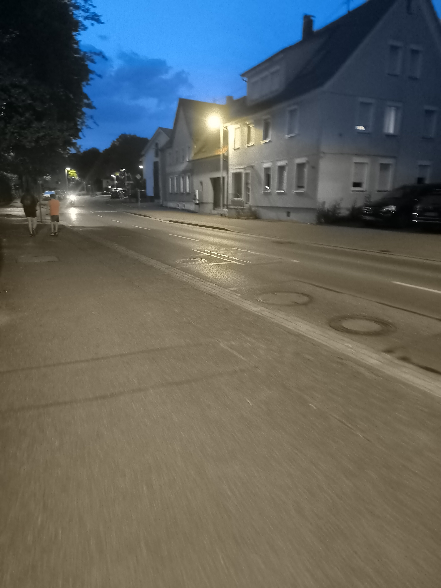 An outdoor, eye-level, wide-angle shot captures a street scene at dusk in an unknown urban location. The sky is a deep blue with some clouds. On the left side of the frame, two individuals are walking on a sidewalk next to a tree. One person is wearing an orange shirt and dark shorts, while the other is wearing a dark top and light-colored shorts. Across the street, several buildings are visible, illuminated by streetlights. A dark-colored car is parked in front of one of the buildings. The road in the foreground has white lane markings and two manhole covers. The overall lighting suggests it is either early evening or late night.
