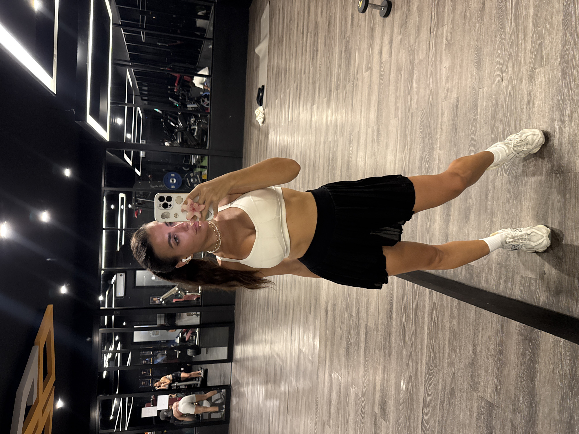 A woman is taking a selfie in what appears to be a gym, based on the presence of exercise equipment visible in the background mirrored walls. She is dressed in a white athletic top and a black pleated skirt, and is wearing white sneakers with white socks. She is holding a cell phone in her right hand, taking a photo of herself. Her hair is pulled back in a low ponytail, and she is wearing a necklace. The gym is well-lit, with dark-colored walls and reflective surfaces. The floor is a light grey color with a wood-grain pattern. The background contains various pieces of workout equipment. The lighting is predominantly artificial. The location is North Kuta, Indonesia.