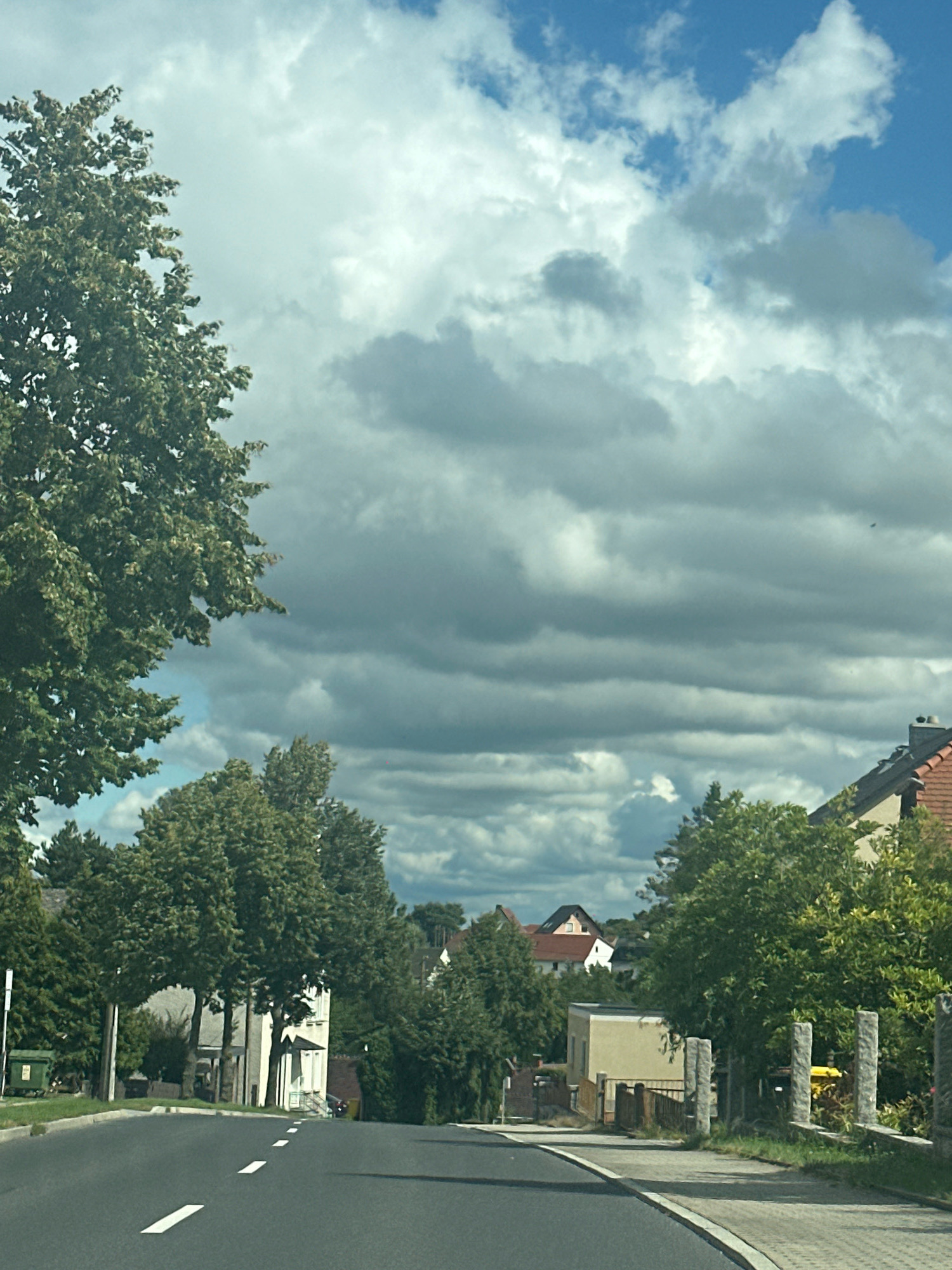 The image depicts a street scene in Panschwitz-Kuckau - Pančicy-Kukow, Germany. The foreground shows a paved road with white dashed lines marking the lanes, curving to the right. On the left side of the road, a large, leafy green tree dominates the frame, with its branches extending towards the sky. Beyond the tree, a white building with a dark roof is visible. Further along the street, several houses with red-tiled roofs and green trees line the sides. In the background, more houses and trees are scattered across a slightly hilly landscape. The sky above is filled with a mix of white and gray clouds, with patches of blue sky showing through. The overall impression is a quiet, suburban street on a partly cloudy day.
