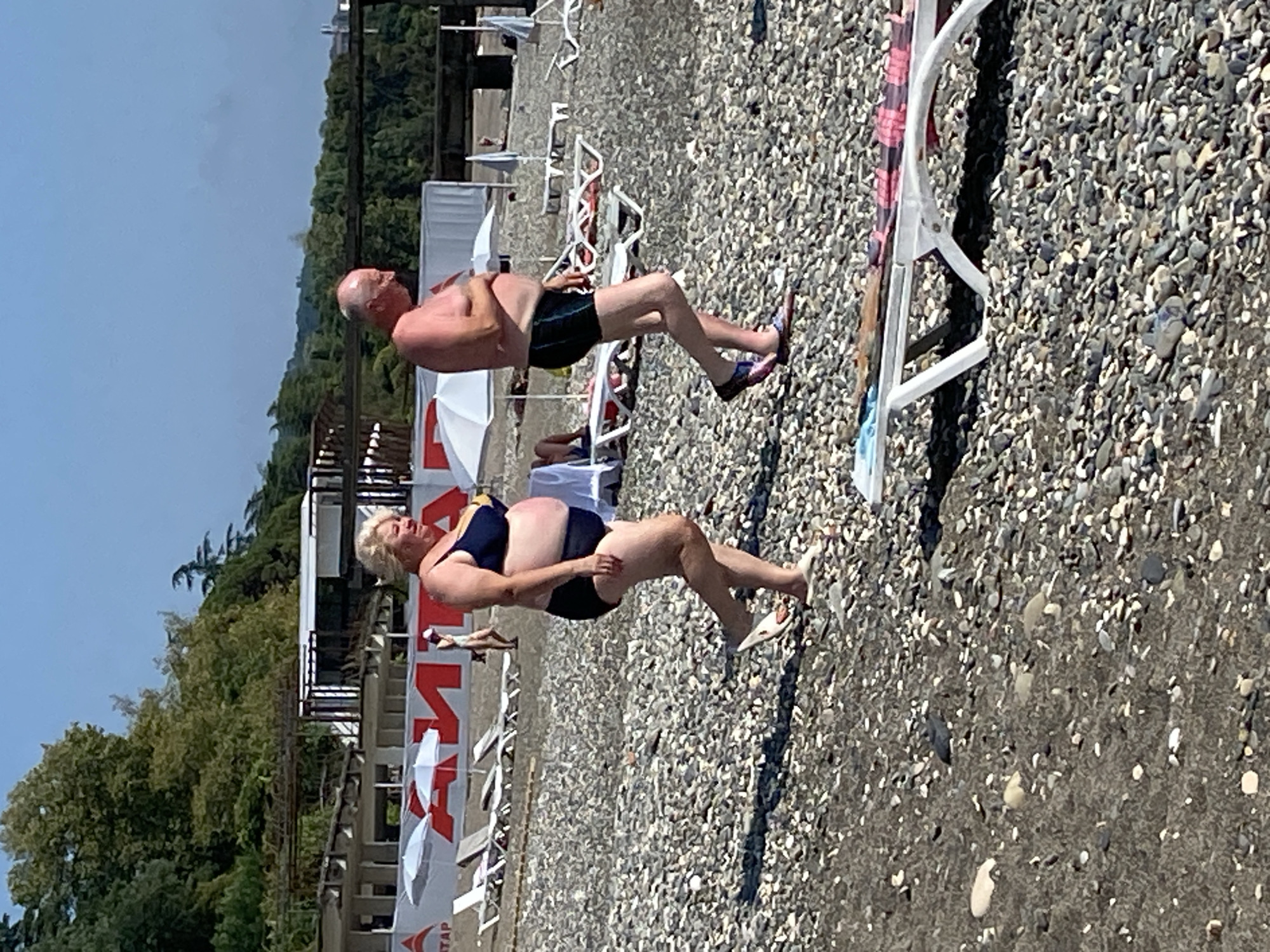 On a pebble beach in Chabkwara, Abkhazia, two individuals walk. On the left, a woman with blonde hair wears a blue bikini and white slides, her body is full-figured. On the right, a bald man with fair skin wears black swim trunks, his body is full-figured. In the background, there is a building with white and red signage, white umbrellas, and lounge chairs. The sky is clear and blue.