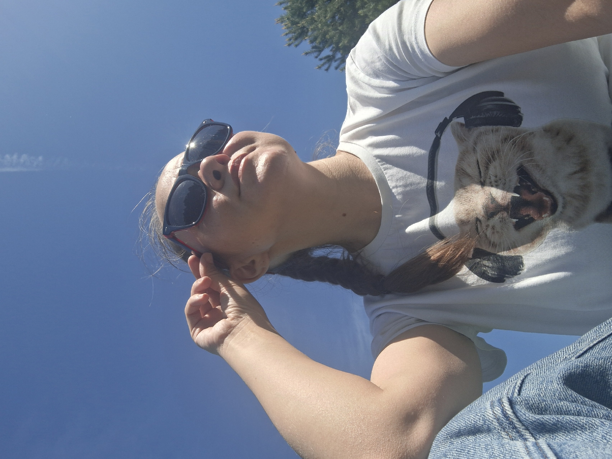 A low-angle shot depicts a person with fair skin wearing sunglasses and a white t-shirt with a cat print, against a clear blue sky with a faint contrail. The person's hair is styled in a braid. The background suggests an outdoor setting with a treetop visible. Clothing details include blue denim jeans. No specific information confirms the location is Chaikovsky, Russia.