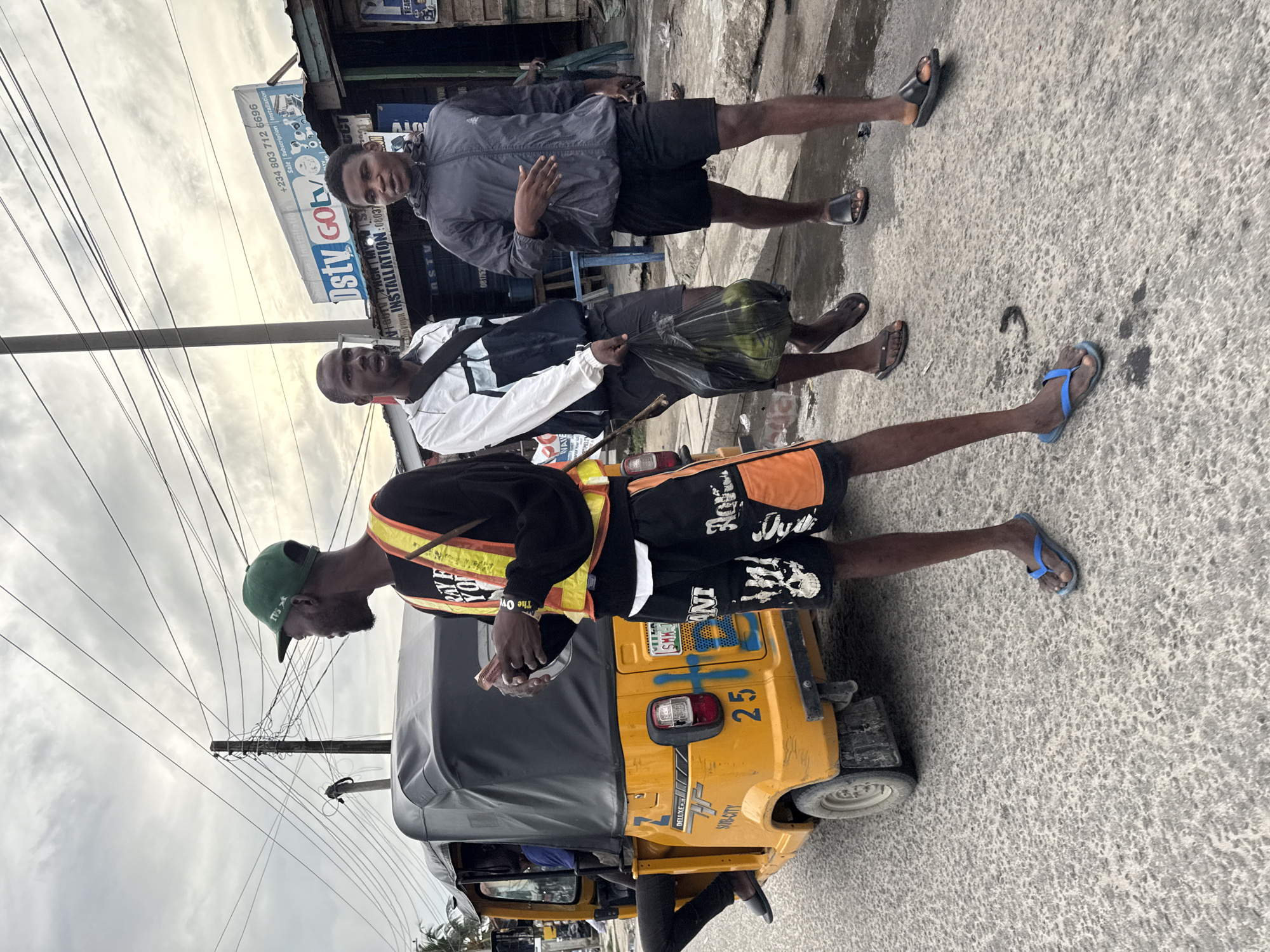 Here is a forensic description of the image:
**Scene Overview:**
The image depicts a street scene in what is likely Ado, Nigeria. The lighting suggests it is daytime with overcast skies. The road is paved and shows wear. A yellow and black "Keke Napep" three-wheeled taxi is parked on the roadside. Three men are positioned near the vehicle.
**Subjects:**
1. A man wearing a black long-sleeved shirt with a yellow and orange reflective vest, black and orange patterned shorts, and blue flip-flops is standing next to the Keke Napep. He is wearing a green baseball cap.
2. A second man wearing a white jacket over a black shirt, with black shorts, and black slides, is carrying a black plastic bag.
3. A third man wearing a grey jacket over a black shirt, black shorts, and black slides stands behind the second man.
**Objects:**
* Keke Napep three-wheeled taxi with "TVS" and "SUB-CITY DELUXE" visible. The rear license plate number is partially visible. "25" is written on the body of the taxi.
* A black plastic bag, likely containing groceries.
* Power poles and overhead electrical wires.
* A commercial signboard "DSTY GOTO ... INSTALLATION: 08037" is visible in the background. "+234 803 712 6696" is also on the sign.
**Actions and Interactions:**
* The man in the reflective vest appears to be interacting with someone inside the Keke Napep, possibly the driver.
* The other two men are standing near each other. The man in the grey jacket is gesturing with his right hand.
**Environment:**
The scene appears to be a commercial area or roadside in Ado, Nigeria, with evidence of businesses nearby.