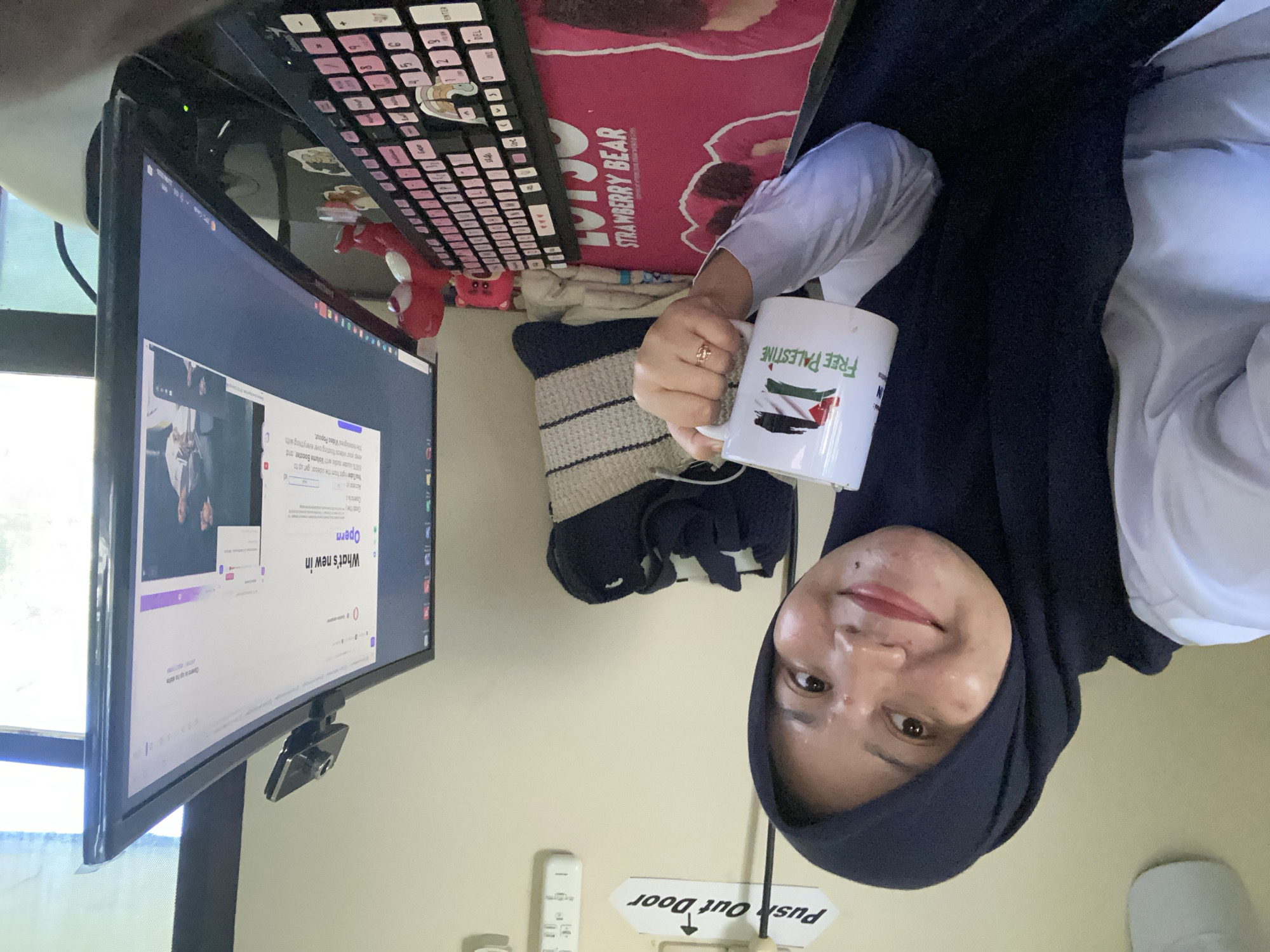 The image shows a woman in a black hijab and white shirt, holding a mug that says "Free Palestine" and features the Palestinian flag. She is seated at a desk in front of a computer monitor, which displays a web page from the Opera browser. A keyboard with pink keycaps and a webcam are visible on the desk. A stuffed toy bear is also on the desk, to the left of the monitor. In the background, a sign on the wall reads "Push Out Door." The environment appears to be an indoor setting, likely an office or home office. There is no specific indication of the city of Banjarbaru, Indonesia in the image.