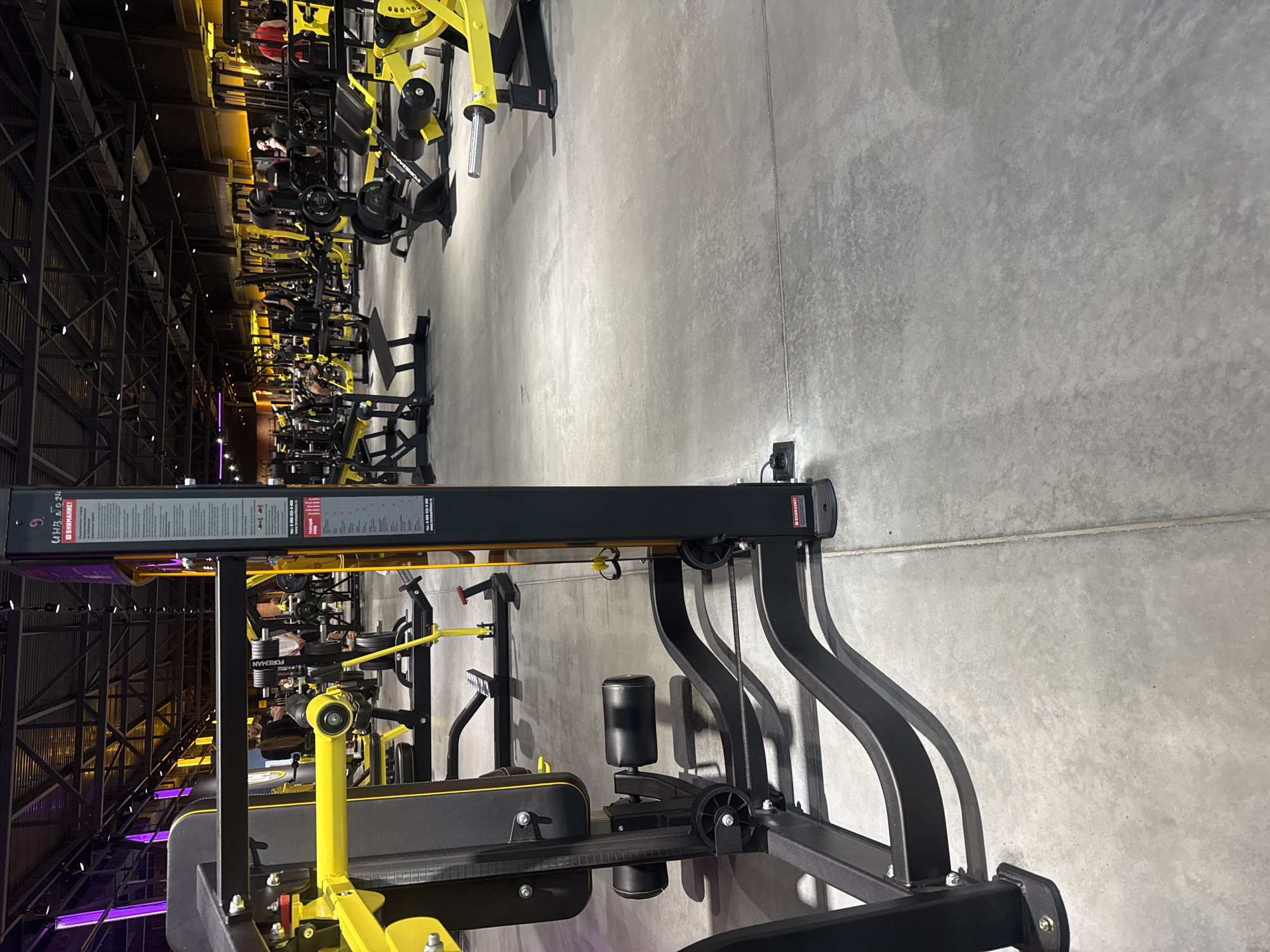 The image shows the interior of a modern, well-lit gym. The floor is concrete, and various weightlifting machines, predominantly yellow and black, are arranged throughout the space. Some of these machines appear to be cable machines and weight benches. In the background, several individuals are visible, some engaging in workouts and others standing around. The gym is decorated with purple and yellow lighting fixtures. The overall environment suggests a commercial fitness facility. The location is identified as Komsomolsk-on-Amur, Russia.