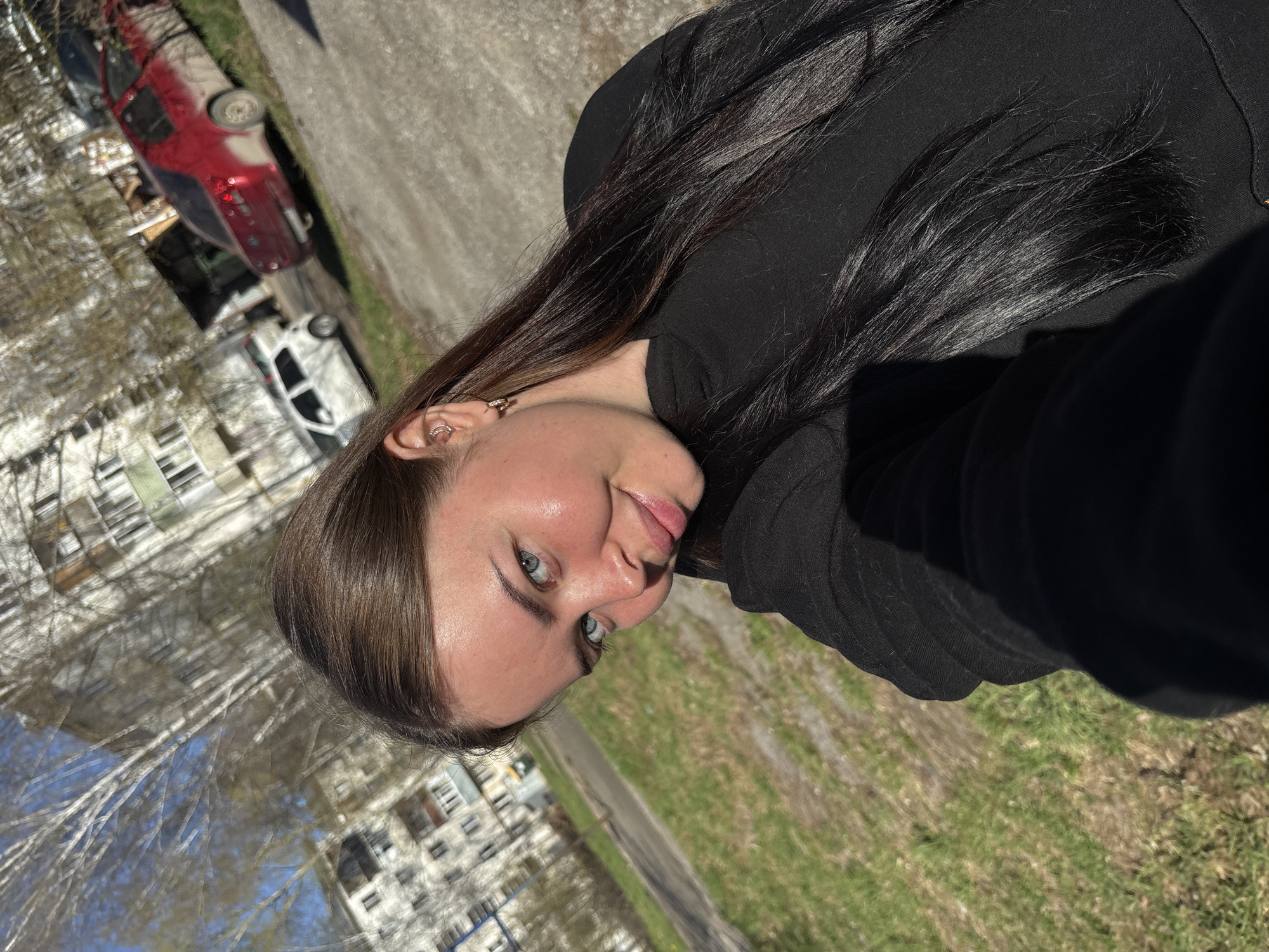A rotated image depicts a light-skinned woman with dark hair, likely in her 20s, wearing a black top. She is looking at the camera. In the background, visible buildings suggest an urban environment. A street is visible, with two vehicles parked along the roadside. A red vehicle appears to be turned over. It is possible the location is Izhevsk, Russia.