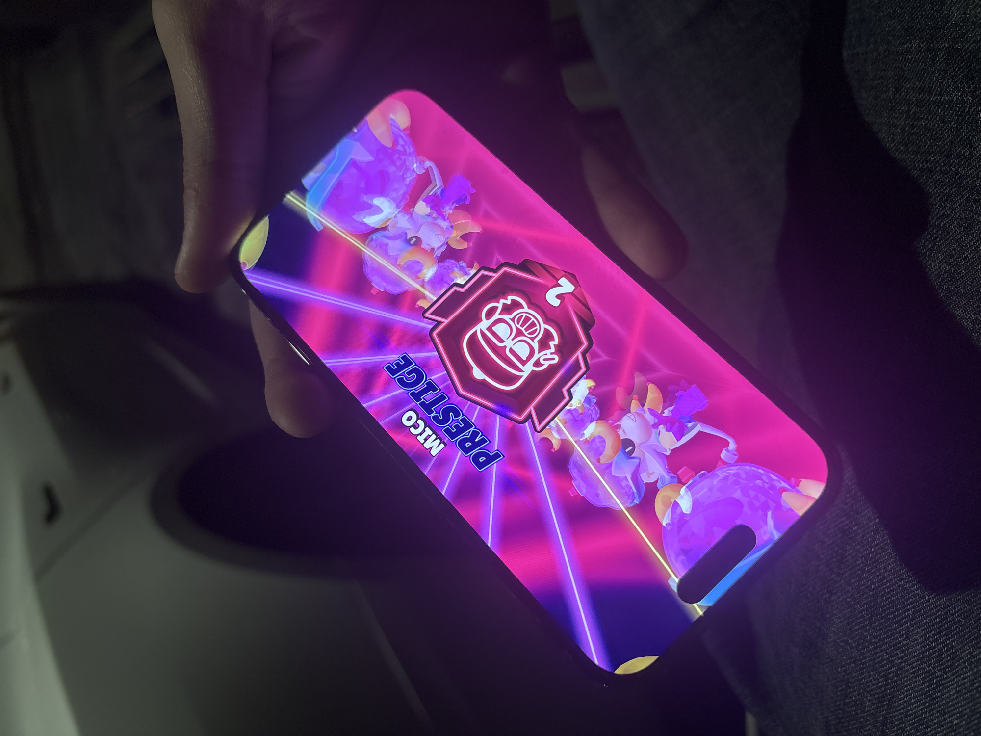 The image shows a hand holding a smartphone displaying a game screen. The game appears to be "Brawl Stars", specifically showing the "Mico Prestige 2" character. The background is a vibrant mix of pink and purple with neon-like streaks.
There is no information in the image to suggest the location is Yerevan, Armenia.
