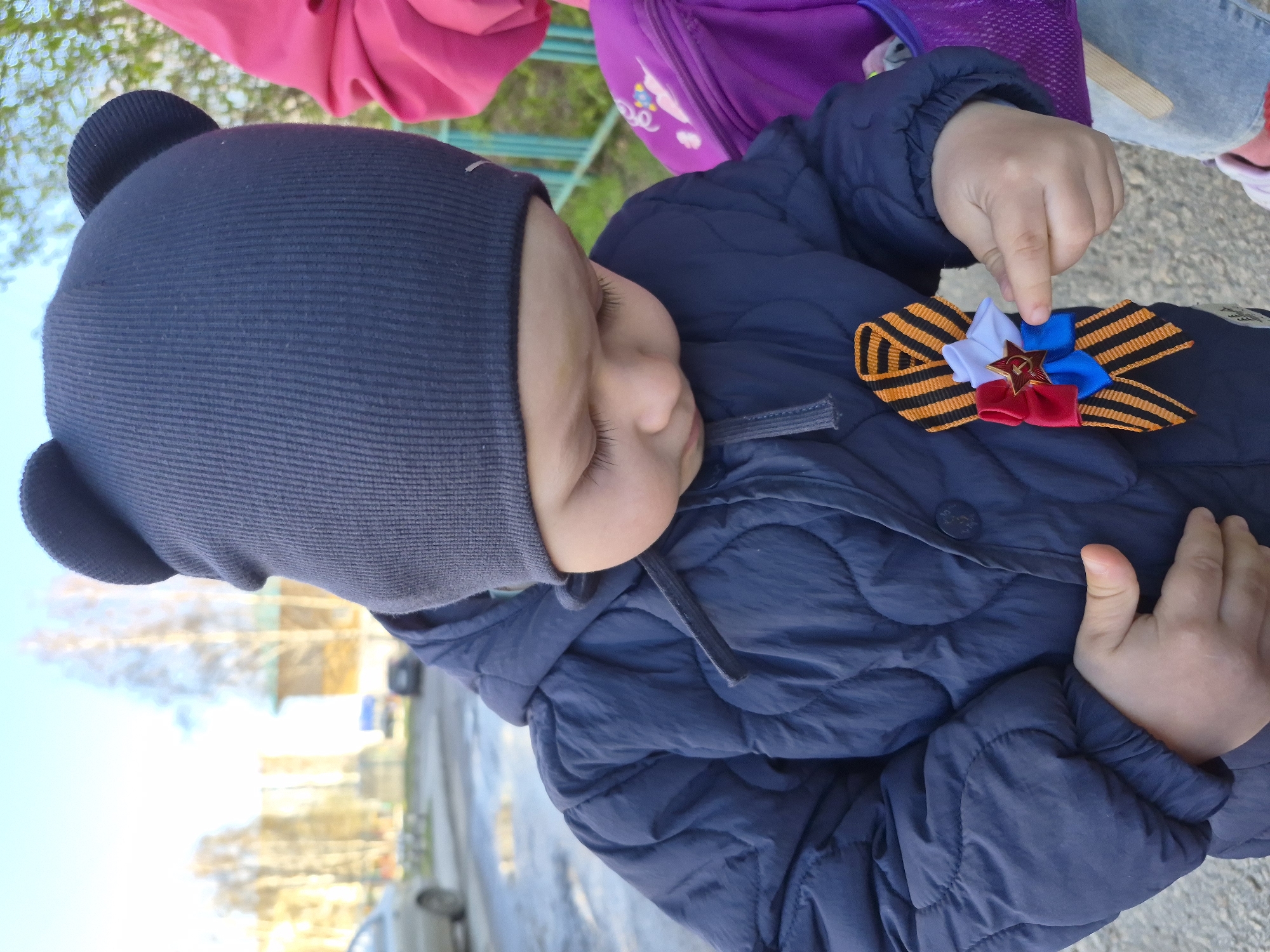 A child with a navy blue winter hat with bear ears is wearing a blue quilted jacket and touching a ribbon pinned to their jacket. The ribbon is orange and black striped with a star, red, blue and white bow. The child is standing outside in what appears to be a city, with buildings and bare trees in the background. The scene appears to be in Tobolsk, Russia, based on the ribbon which is a symbol of Victory Day in Russia.