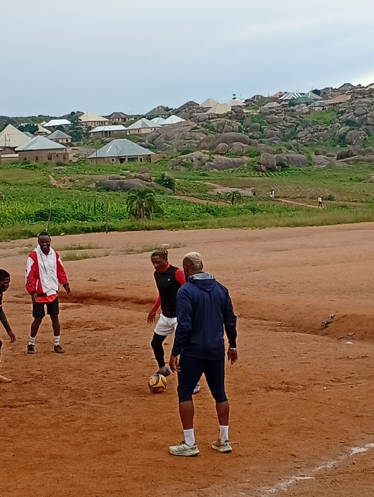 The image captures an outdoor scene in the city of Jos, Nigeria, where a group of young men are playing soccer on a dusty, reddish-brown field. The foreground is dominated by the playing area, with the men in various athletic attire. One man in the center, wearing a black sleeveless top and white shorts, is actively engaged with the soccer ball at his feet. To his left, a man in a red and white jacket and black shorts appears to be watching the game. Another individual, seen from behind and wearing a navy blue hooded jacket and dark shorts, stands with his back to the camera, also seemingly observing or participating.
In the midground, the terrain transitions into a lush green, grassy area interspersed with scattered vegetation and some palm trees. Beyond this, the background reveals a distinctive landscape of rocky hills characteristic of Jos. Numerous buildings with corrugated metal roofs are scattered across these hillsides, suggesting a residential area nestled within the rugged terrain. The sky above is overcast, casting a soft, diffused light over the entire scene. The presence of both the playing field and the hilly, residential background firmly places this activity within the urban landscape of Jos.