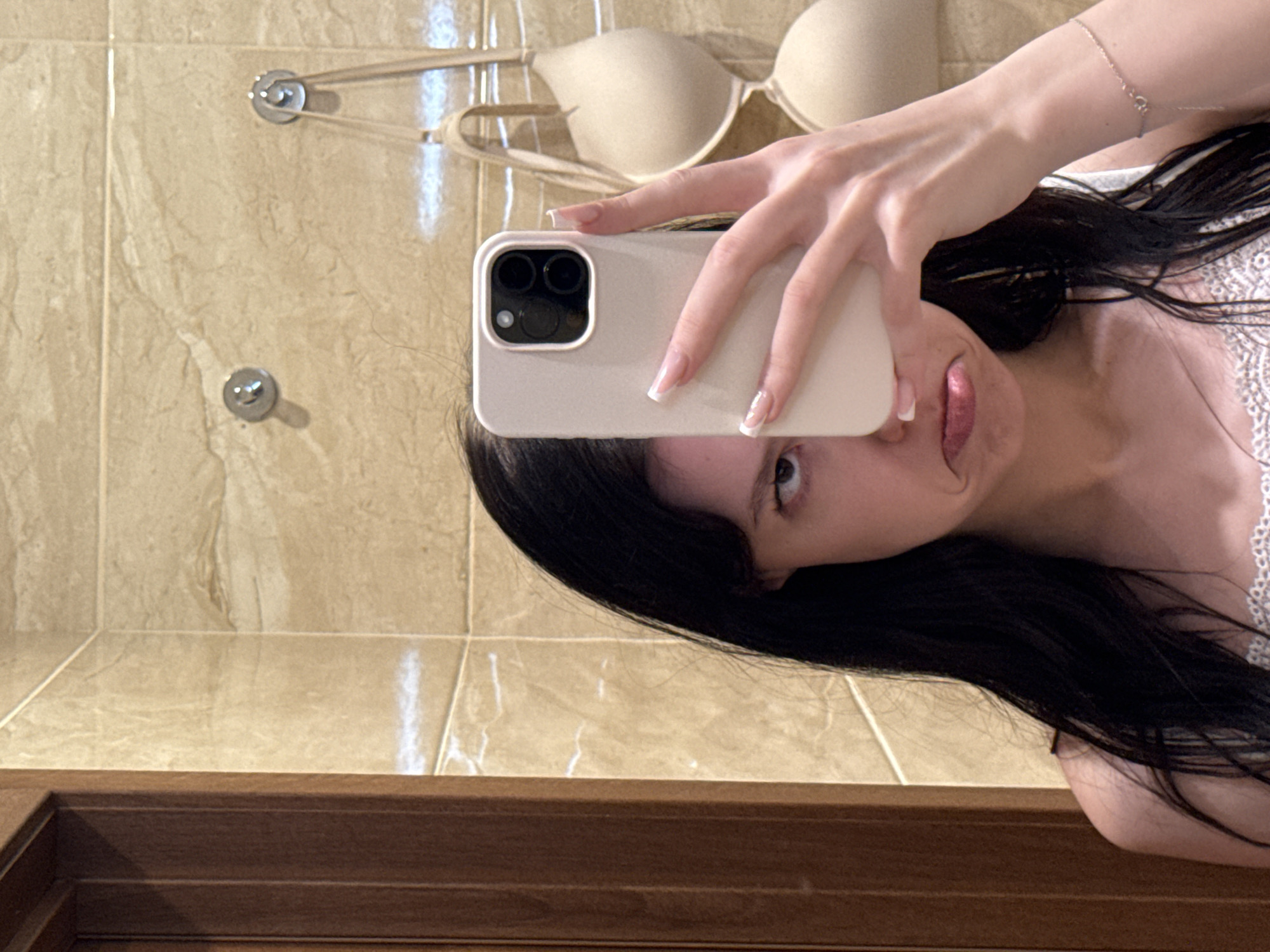 In a bathroom with beige tiled walls, a young woman with long, dark hair takes a selfie with her phone, which is held up to cover half of her face. She is wearing a white lacy camisole and has a pouty expression. Behind her, a beige bra is hanging on a hook. A wooden door frame is visible on the left side of the image. The lighting suggests the photo was taken indoors, possibly in an apartment or hotel bathroom. The presence of bathroom tiles, a door frame, and personal items like a bra indicate a private domestic setting. The overall scene is consistent with a typical bathroom environment.