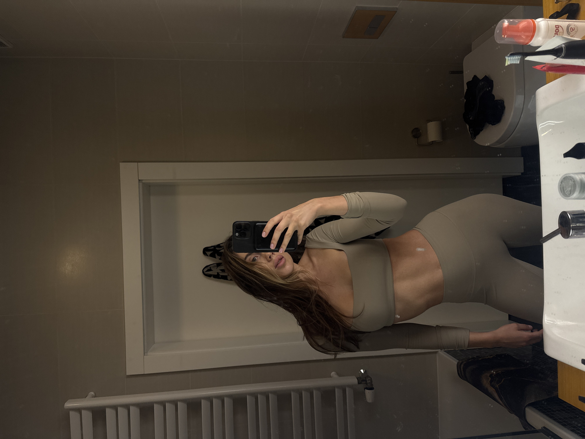 A woman is taking a selfie in a bathroom. She is wearing a matching set of leggings and a long-sleeved crop top. The bathroom has beige tiles, a white sink, a toilet, and a radiator. There is also a door with a rack of rabbit ears on the other side of it. The woman is holding a phone in her hand, and her hair is long and brown. The location is assumed to be in Warsaw, Poland, but there is no evidence of this.