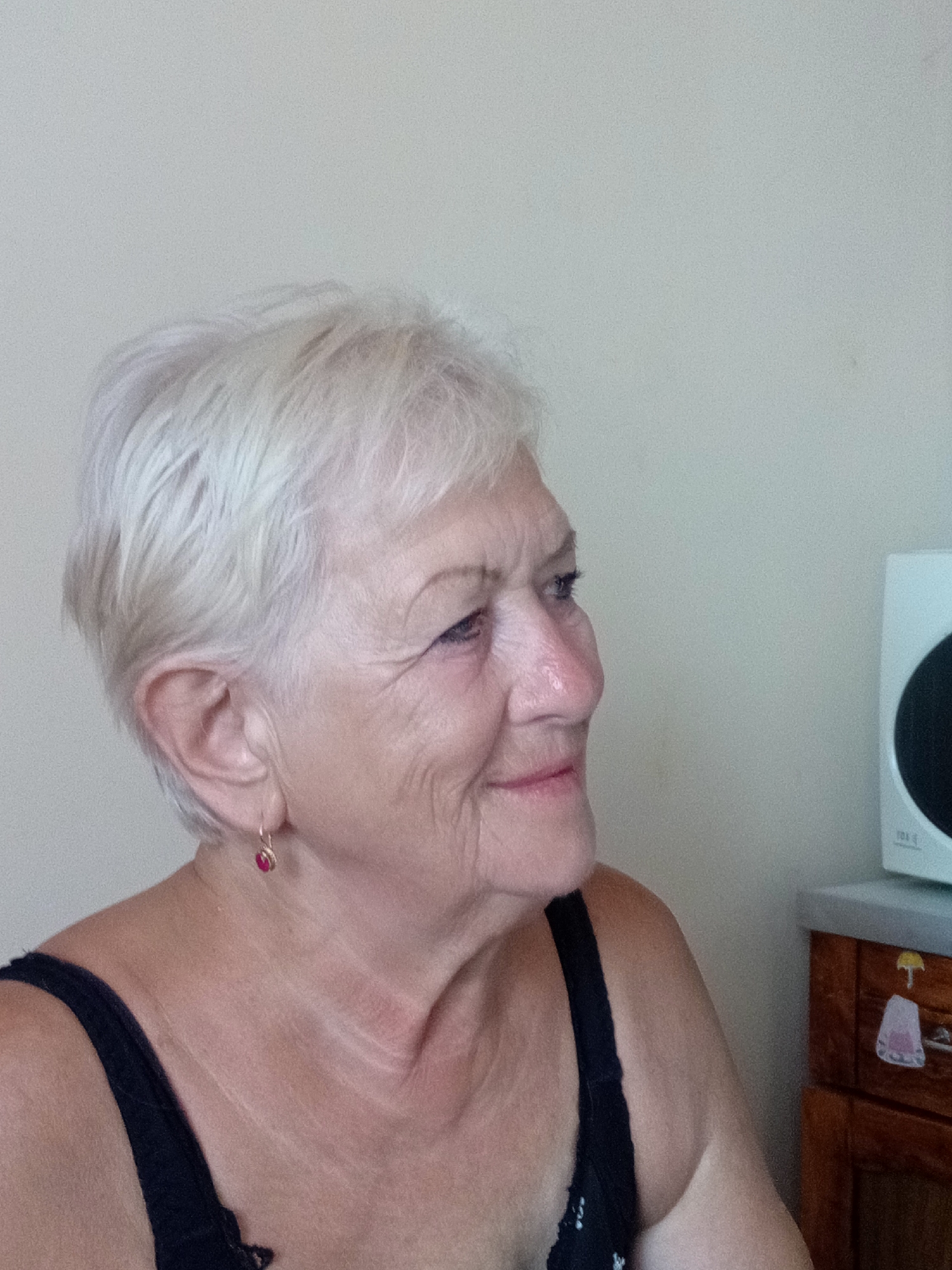 The image shows a close-up, profile view of an elderly woman with short, blonde hair and fair skin. She is wearing a black top. She has a small, red earring in her left ear. Her facial expression is neutral, and she appears to be looking to her right. The background is a plain, light-colored wall. To the right of the woman, a white object with a black circular element is visible, and below that, a portion of a wooden cabinet with some decorative items is present. The scene appears to be indoors. There is no indication in the image that it is located in Tiachiv, Ukraine.