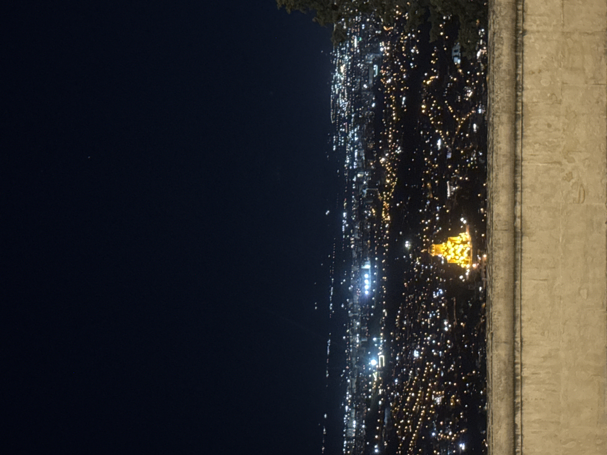The image shows a nighttime panoramic view of Tbilisi, Georgia, as seen from a high vantage point, likely a viewpoint or a hilltop. In the foreground, a textured, light-colored concrete wall or railing runs horizontally across the bottom of the frame, suggesting it's part of a balcony or viewing platform. Beyond the wall, the city lights of Tbilisi stretch across the landscape, creating a dense, sparkling tapestry of warm yellow and white lights. The lights suggest a sprawling urban environment with numerous buildings and streets.
Prominently featured in the middle ground, to the right of the center, is a brightly illuminated structure, possibly a church or a significant building, bathed in a golden yellow light. Its architecture appears to be distinctive, with multiple levels and a conical or domed roof. The glow from this structure is quite strong, making it a focal point within the city lights.
The sky above is a deep, dark navy blue, indicating it is nighttime. A few faint, distant lights or celestial bodies are barely visible in the sky. To the far right edge of the image, the dark silhouette of a tree or foliage can be seen, partially obscuring the view of the city. The overall impression is one of a vibrant city at night, viewed from a serene overlook. There are no people or animals visible in the scene.