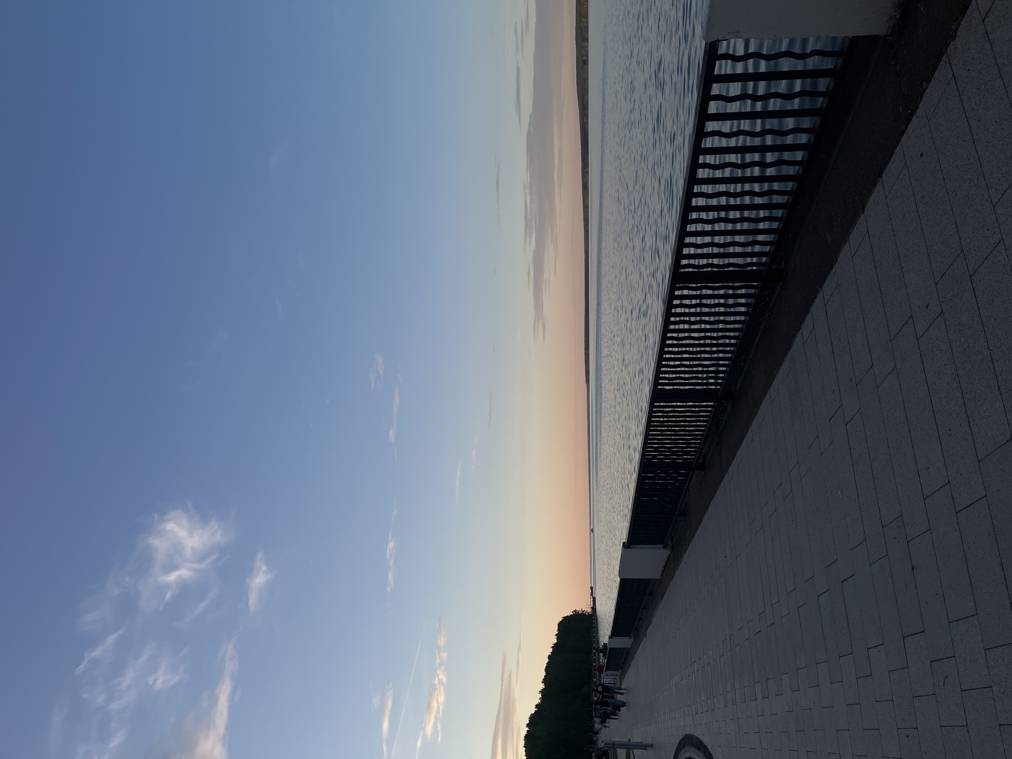 The image captures a panoramic view of a waterfront promenade in Chaikovsky, Russia, during twilight. The scene is dominated by a vast body of water, likely the Kama Reservoir, stretching horizontally across the midground. The sky above transitions from a clear, deep blue in the upper right to softer pastel hues of pink and orange near the horizon on the left, indicative of sunset or sunrise. Scattered, wispy cirrus clouds are visible in the upper left quadrant of the sky.
In the foreground, a paved promenade composed of grey rectangular tiles extends from the lower left towards the distant horizon on the left. A dark, ornate metal railing, featuring vertical, undulating bars, separates the promenade from the water, supported by periodic white concrete blocks. Several indistinct human figures are walking along the promenade on the far left, appearing as silhouettes against the diminishing light.
To the far left, a dense line of dark green trees forms a forested bank, receding into the distance along the water's edge. Across the water, a distant, low-lying landmass, also appearing forested, forms the opposite bank on the horizon, extending from the center to the right. The water surface is calm, reflecting the sky's muted colors and showing subtle ripples. A small, dark object, possibly a boat, is discernible on the water near the forested bank on the left horizon. The overall lighting casts the foreground elements into shadow while illuminating the sky and water with soft, diffused light.