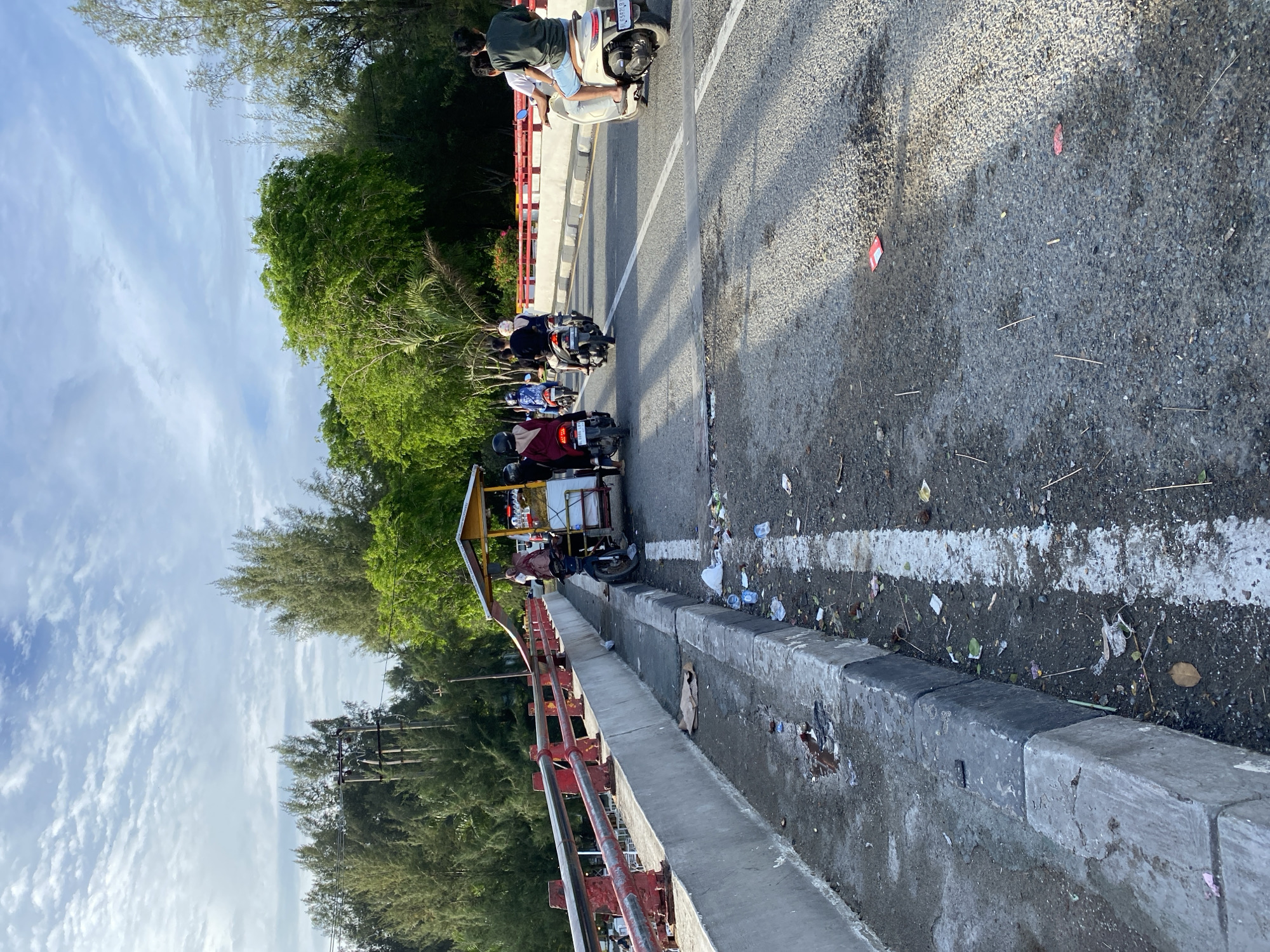 The image depicts a street scene on a bridge in Banda Aceh, Indonesia. Several people are riding motorbikes, with some carrying passengers. One motorbike is attached to a cart that appears to be a mobile food stall, with a person standing next to it. The bridge has a concrete barrier with red railings. The roadway is asphalt and shows signs of wear and tear, with debris and litter scattered along the side and on the road. The surrounding environment includes lush green trees and a cloudy sky. Power lines and poles are visible in the background. The lighting suggests it is daytime, possibly late afternoon due to the long shadows.