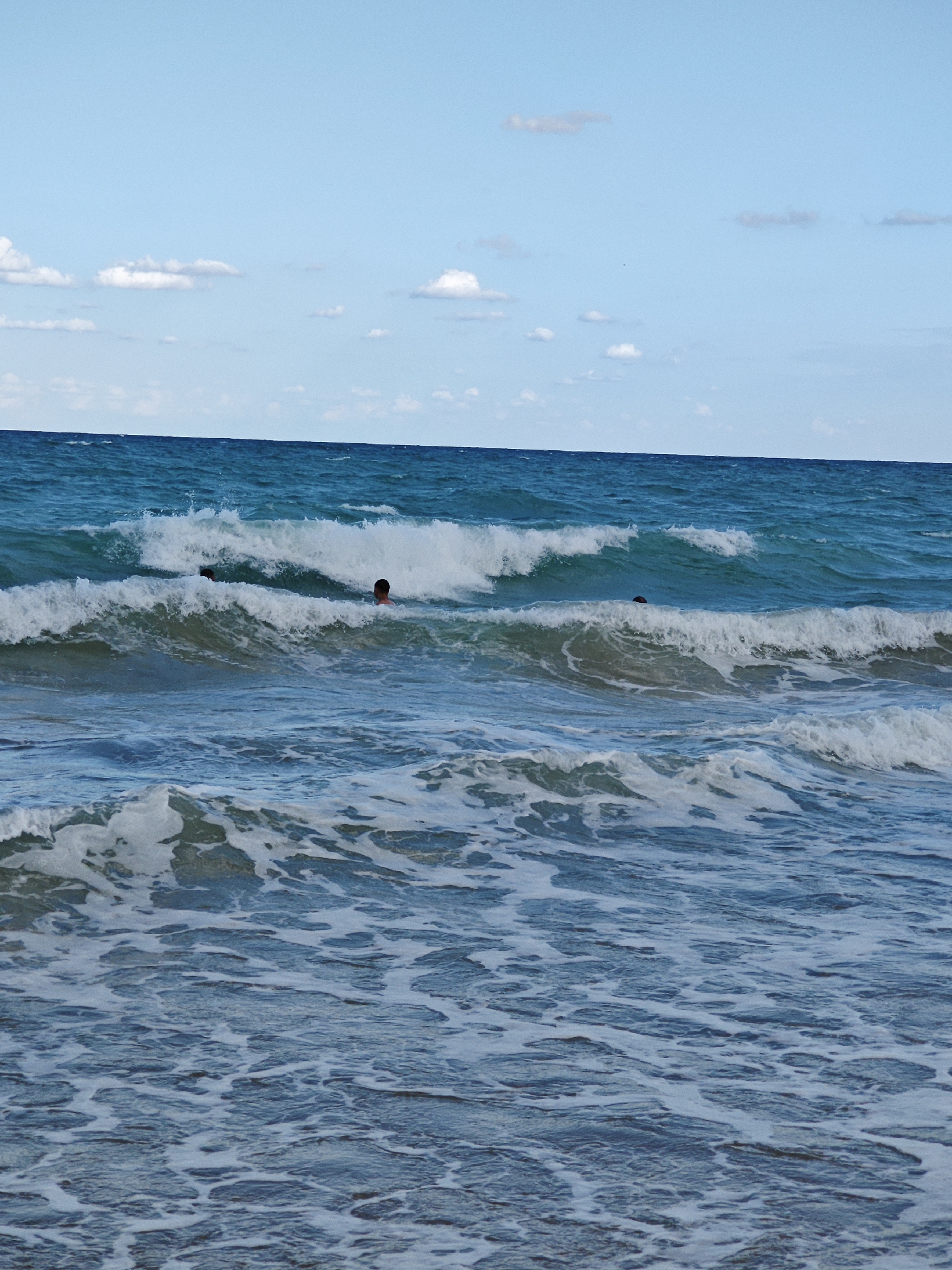 The image shows two people swimming in the ocean. The water is a deep blue with whitecaps from the waves. The sky is clear with a few white clouds. The scene appears to be a beach on a sunny day. The people are in the water, with their heads visible above the surface. They are swimming and playing in the waves. The overall impression is one of a fun and relaxing day at the beach.