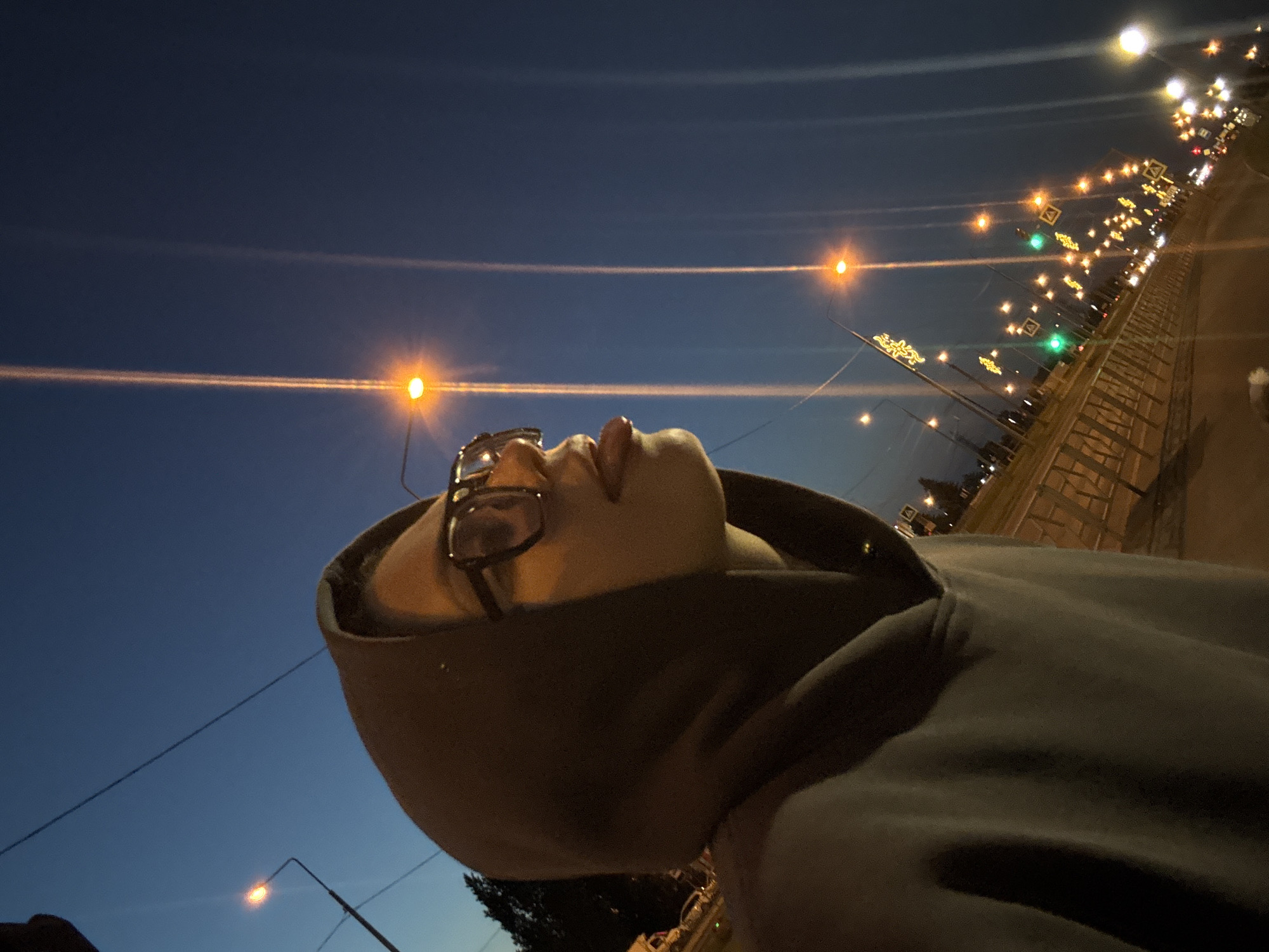 The image depicts an individual in a dark hooded sweatshirt and eyeglasses, shown in profile, facing right, with their tongue slightly extended. The scene is set at dusk or night in an urban environment in an unknown city and country. The person's face is illuminated by warm-toned artificial light. In the background, a street or highway stretches into the distance, lined with numerous streetlights that emit prominent vertical light streaks due to lens flare. Some streetlights feature glowing yellow decorative elements. Green traffic lights and pedestrian crossing signs are visible along the road, alongside a metal road barrier. Distant vehicle lights suggest ongoing traffic. The sky is a deep blue, indicative of twilight or night.