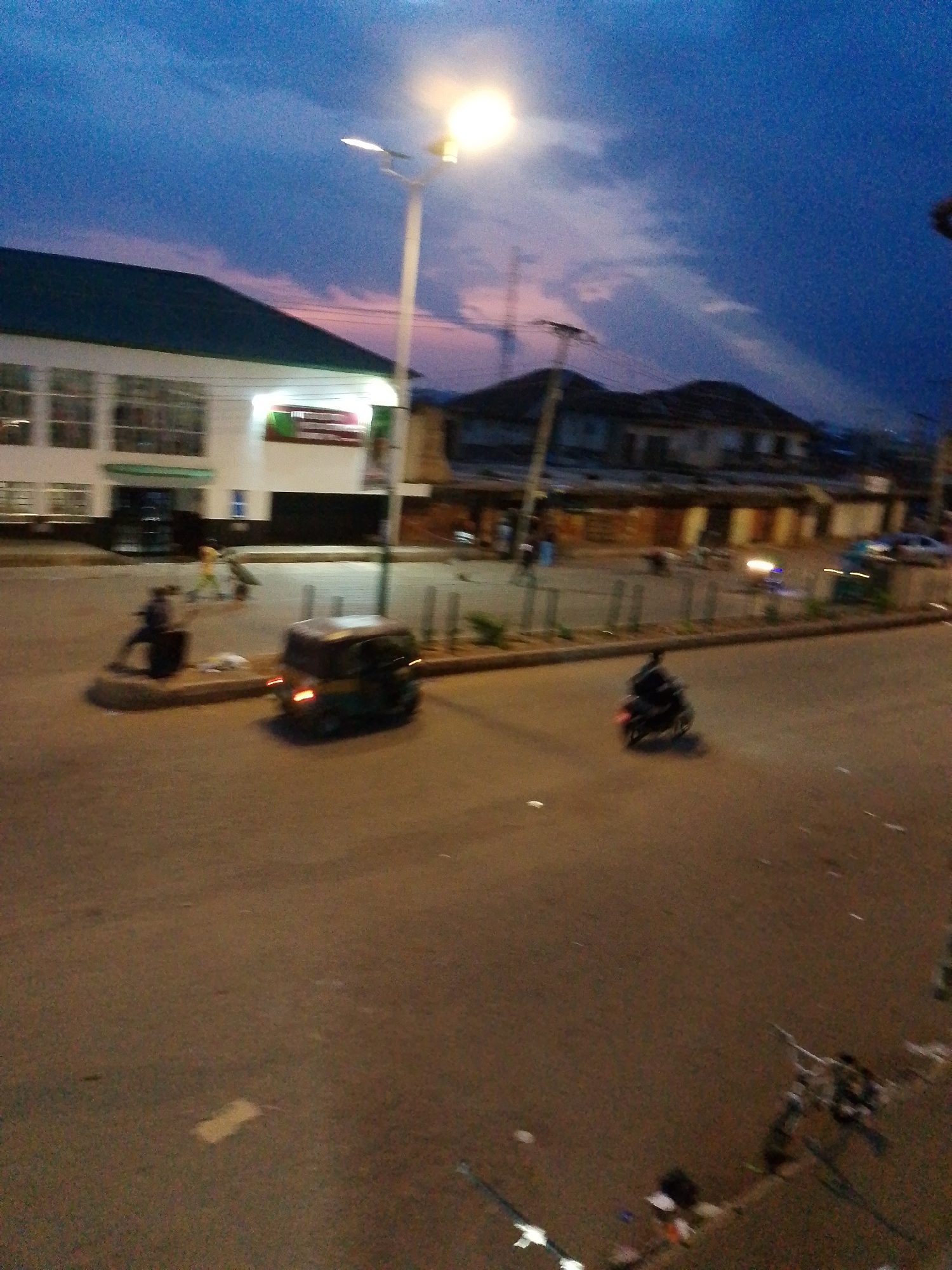 At dusk in Bukuru, Nigeria, an intersection is captured. The sky displays a gradient of deep blue to purple hues. A streetlamp illuminates a portion of the scene from the upper right. On the left, a white building with a green roof features multiple windows and a sign. Across the street, older buildings with visible roofs line the road.
The paved road shows signs of traffic. A yellow and black three-wheeled auto-rickshaw is parked near the center, its rear brake light illuminated. To the right of the auto-rickshaw, a person on a dark motorcycle is moving away from the camera. Further down the street, a few people are visible near the buildings. The ground is littered with debris.