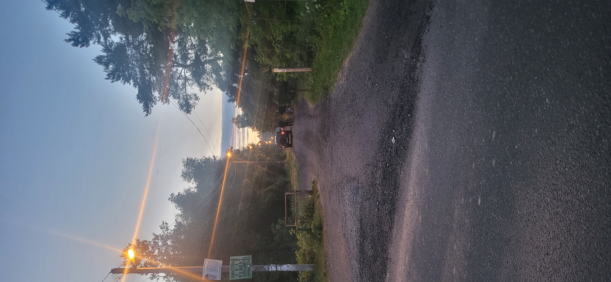 The image depicts a paved road leading into a wooded area at dusk. The sky is a pale blue with hints of orange from the setting sun. Several streetlights are illuminated along the road, casting a warm glow. On the left side of the road, a utility pole stands with two signs attached; one appears to be a portrait and the other has Russian text. A rusty metal gate is partially visible behind the pole. In the distance, a dark-colored SUV is parked on the road, with two figures standing beside it, one holding a bicycle. The surrounding environment is lush with green trees and foliage. The road surface transitions from asphalt in the foreground to a gravelly texture further up.