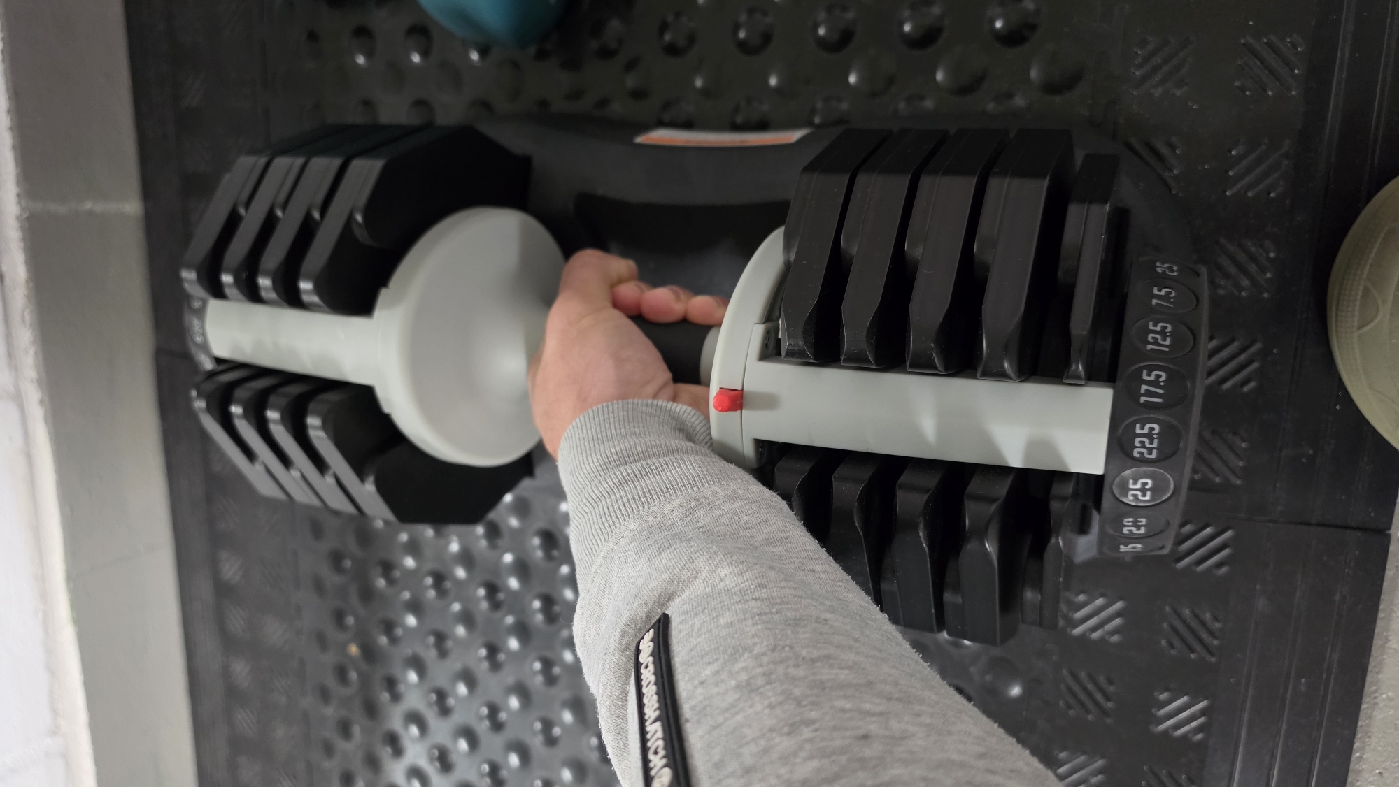 The image shows a person's arm and hand holding the handle of an adjustable dumbbell in a weight rack. The dumbbell is black and grey with weight markings in kilograms on the side. The person is wearing a grey long-sleeved shirt. The rack appears to be on a black rubber mat. The location, Ihringshausen, Germany, is not discernible from the visual information.