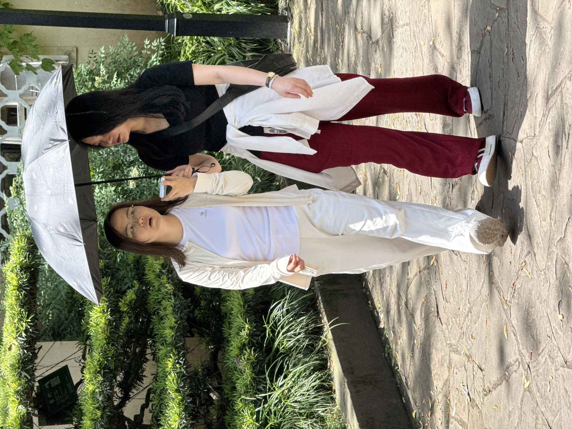 Two women are walking side-by-side under a black umbrella decorated with small yellow designs, located outdoors. The woman closer to the camera is wearing glasses, a white t-shirt, white pants, and a white jacket. The other woman is wearing a black top and dark red pants. A white jacket is draped over her shoulders. The setting appears to be a public area in a city, with greenery visible. It's in Yuzhong District, China. They are walking on a stone path.