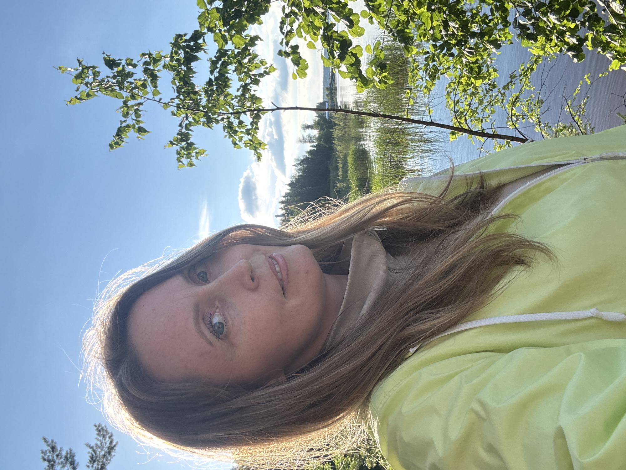 A woman with shoulder-length blonde hair, wearing a light yellow jacket, is taking a selfie outdoors. She has green eyes and is smiling faintly. The background features a lake with reeds and trees, under a bright blue sky with some clouds. The sun is shining from the left, casting a warm glow on her face and hair. The scene appears to be a natural setting, possibly a park or lakeside area, given the presence of water and vegetation. No specific landmarks that would indicate the city of Chaikovsky, Russia are visible.