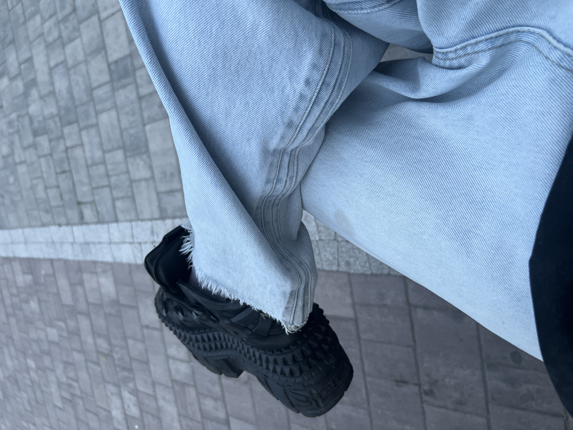 A low-angle, close-up photograph, taken in Saint Petersburg, Russia, depicts the lower legs and feet of an individual from an elevated perspective. The subject is wearing light-wash denim jeans; the left pant leg features a distinctly frayed, unfinished hem. The denim fabric displays its characteristic diagonal weave pattern, along with various creases and folds. Several small, dark, irregular spots are visible on the upper right pant leg. A black athletic shoe with a robust, heavily textured sole, composed of numerous pyramidal protrusions, is worn on the left foot. The background consists of a grey paved surface, likely a sidewalk or pedestrian area, constructed from rectangular paving stones laid in a uniform pattern. A sharp, white linear marking divides the paved surface, running vertically through the scene.
