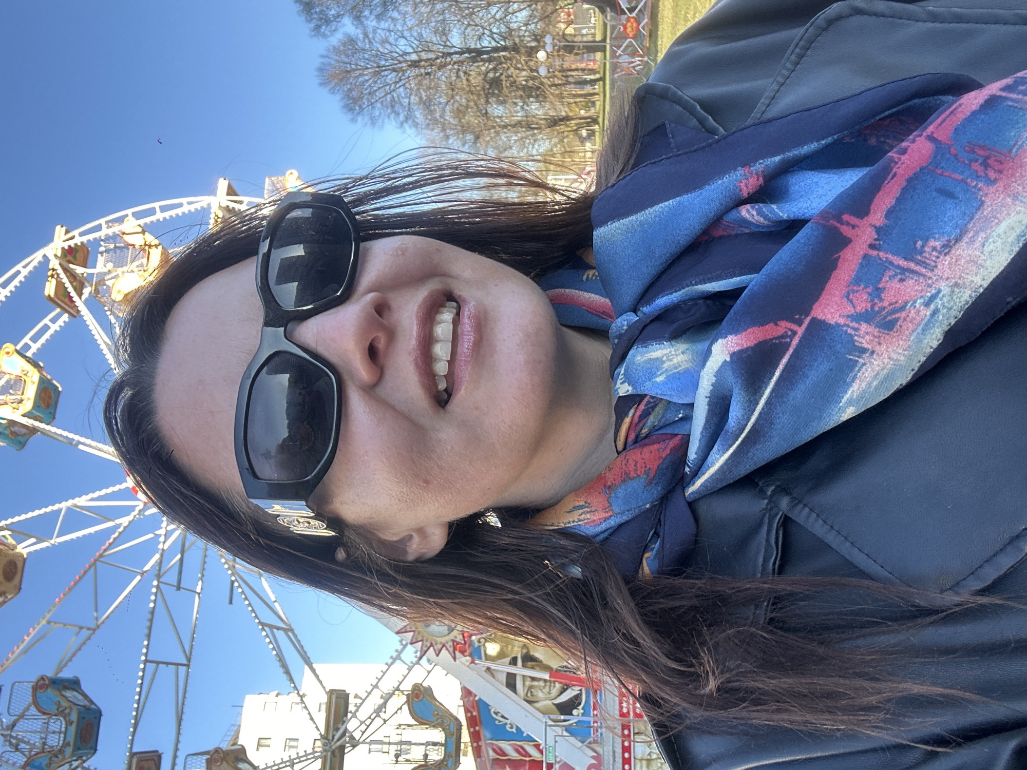 A selfie-style portrait captures a smiling woman at an amusement park in Riga, Latvia, on a bright, sunny day. The woman, appearing to be in her 30s or 40s, has long dark hair and is wearing dark, rectangular sunglasses. Her wide smile reveals her teeth, which appear to have clear aligners or a retainer. She is dressed in a dark blue or black leather-like jacket and a vibrantly patterned scarf with hues of blue, red, and white.
The background prominently features a large Ferris wheel with white spokes and yellow and light blue cabins, set against a clear, deep blue sky. In the distance, bare trees suggest a late winter or early spring setting. Other amusement park rides are partially visible, including one with distinctive red, white, and blue colors and a decorative figure resembling a strongman or king. Another ride features gold and red accents. A small, distant purple object, possibly a kite or small drone, is visible in the sky, as well as a flock of tiny birds. The woman's joyful expression indicates a pleasant and enjoyable experience at the park. No discernible text is visible on any objects or rides.