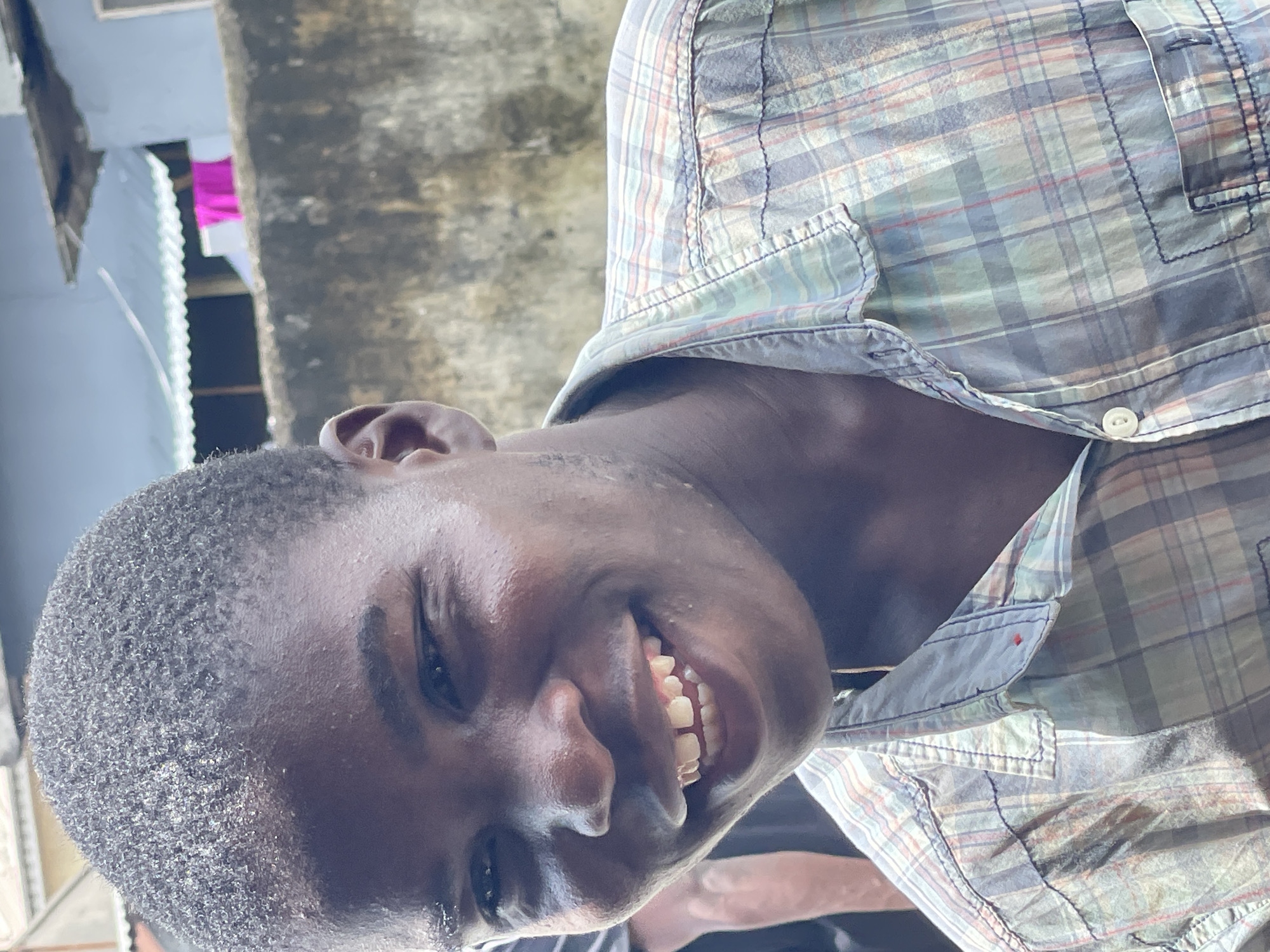 The image is a close-up, rotated portrait of a smiling young man. He is wearing a plaid collared shirt. The background shows an exterior urban scene, with a building fragment on the left, and a wall or pillar on the right. A colorful piece of clothing is hanging in the background. Given the location constraint to Ikorodu, Nigeria, the environment is likely consistent with the urban environment of Ikorodu.