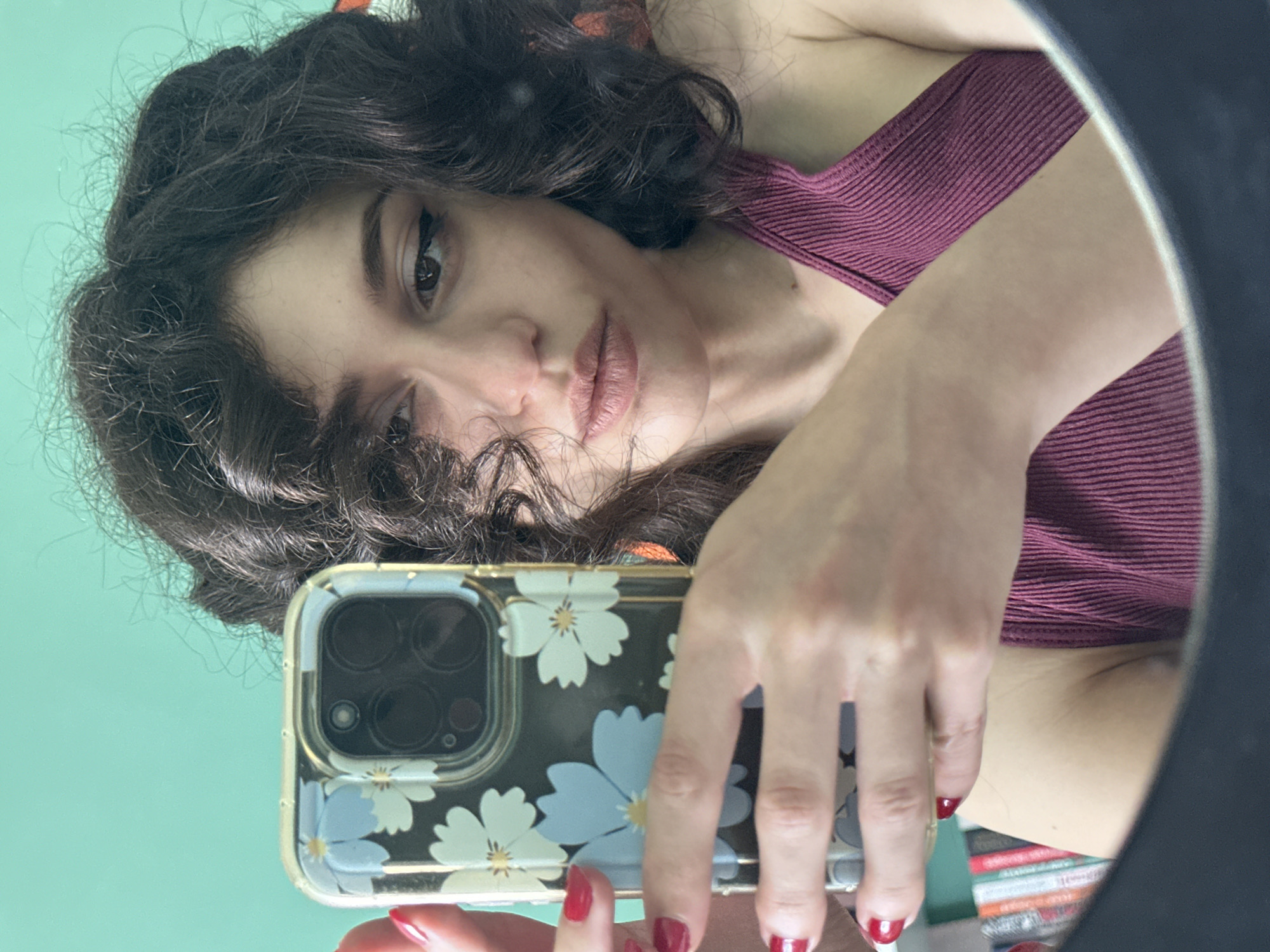 The media file is a portrait-oriented selfie taken in Sofia, Bulgaria. The subject is a young woman with dark, curly hair and brown eyes. She is looking at the camera, her expression is neutral. She is wearing a dark pink ribbed tank top. The background is a light teal wall.
The woman is holding a smartphone with a floral-patterned case in her right hand, which is angled to capture her reflection in a circular mirror. The phone has a multi-lens camera system. Her fingernails are painted red. The image appears to have been taken indoors, possibly in a bedroom or dressing area, with artificial lighting. There is no indication of the time of day or weather. The overall mood is casual and personal.