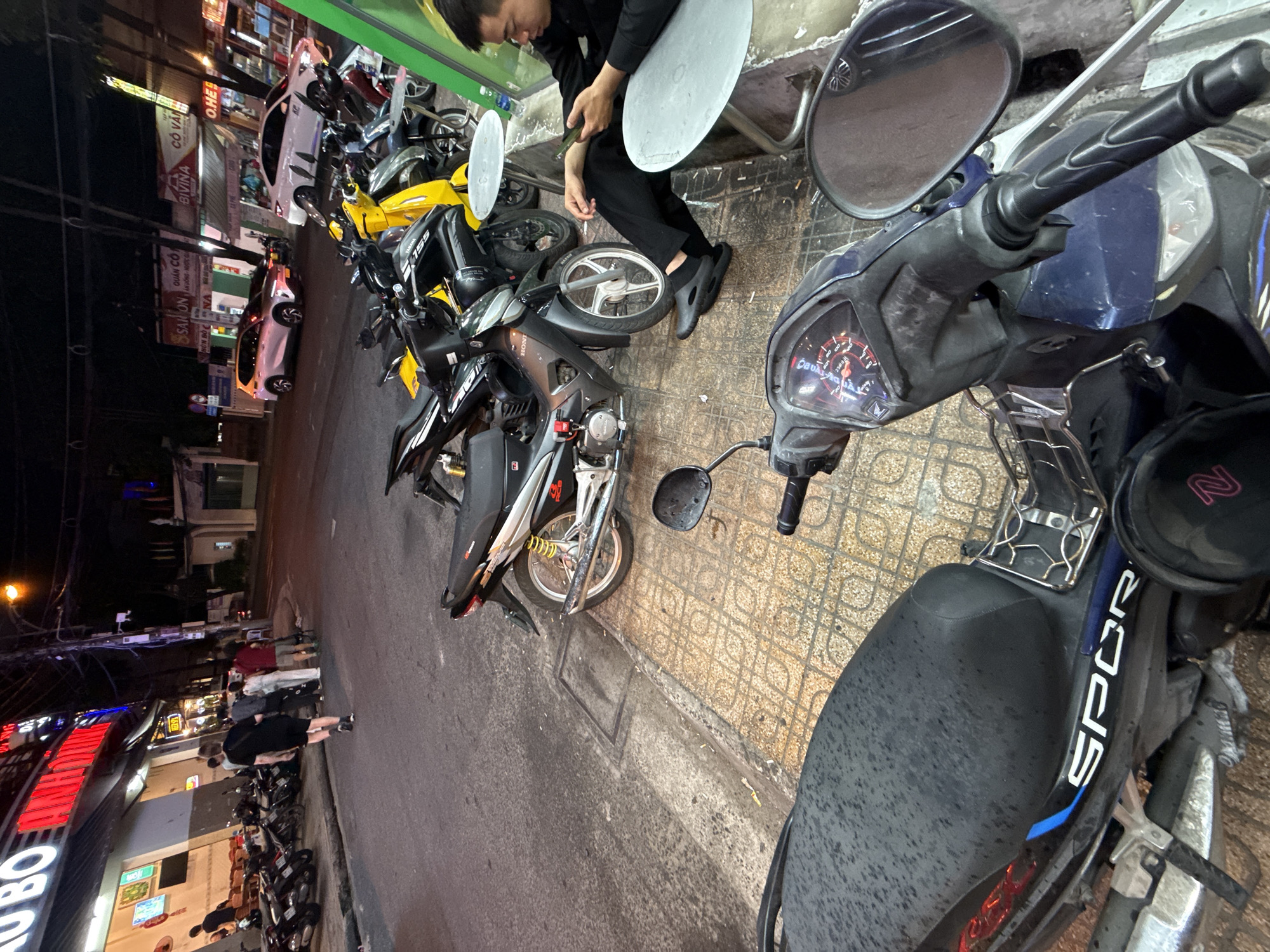 The image depicts a street scene at night in Nha Trang, Vietnam. A row of motorcycles is parked along the sidewalk in front of shops. A man in a black shirt and shorts is walking away from the camera on the street. In the foreground, another motorcycle is parked, and a man in black clothing is sitting on a small table with a mobile phone. The street is illuminated by streetlights and neon signs.