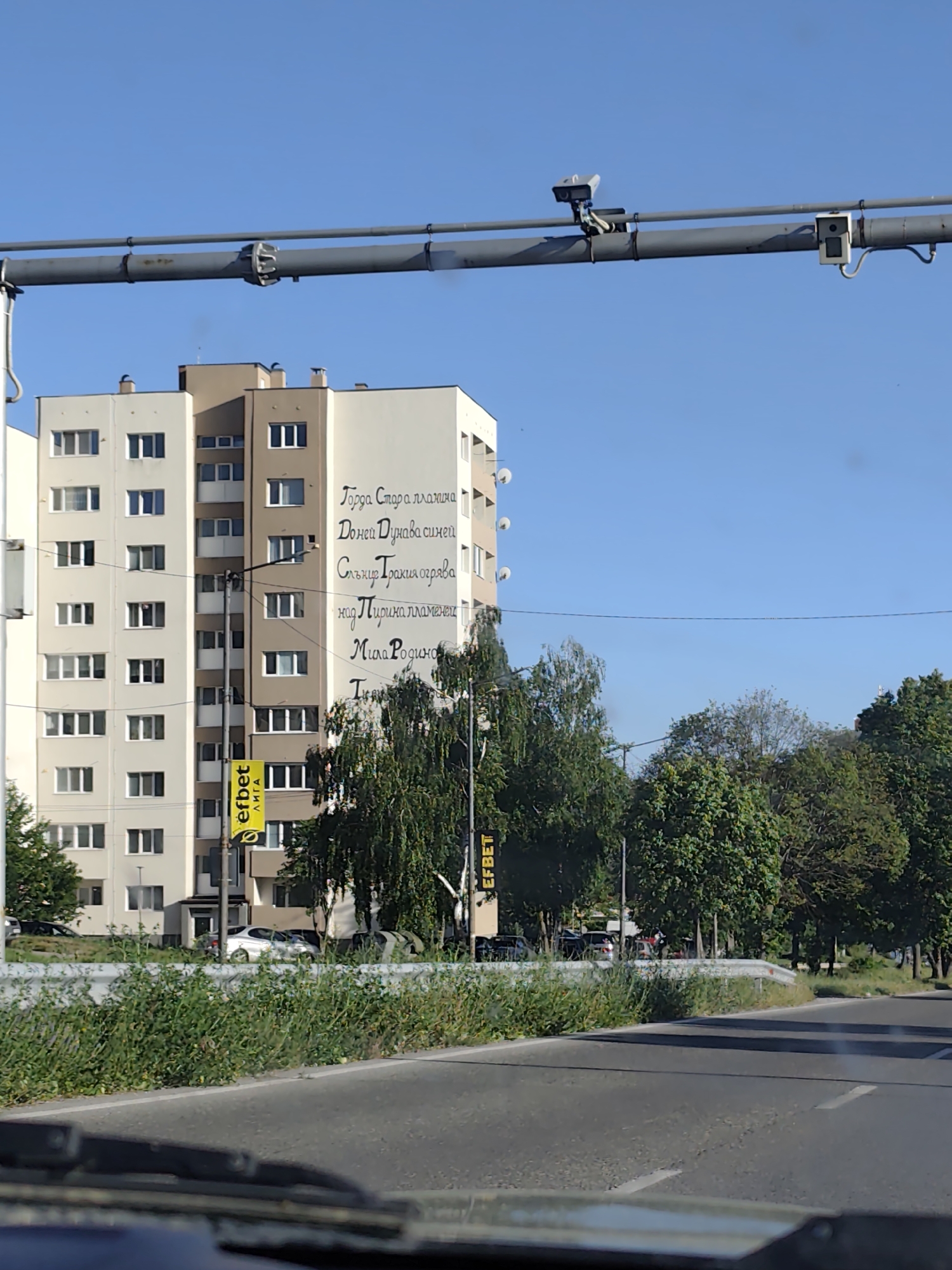 The image shows a street scene from inside a vehicle, possibly a car. In the background is a tall apartment building with beige and white sections. The side of the building has text written vertically in a Cyrillic alphabet; next to it stands a yellow sign with "EFBET ЛИГА" printed vertically. There is also another vertical sign that reads "EFBET" at the side of the road. A variety of green trees are visible next to the building and along the road. Above the street, a metal pipe structure holds surveillance cameras and another unidentified device. The road is mostly empty, with some green foliage and barriers along the edge. The vehicle's dashboard and a slightly out-of-focus windshield are in the foreground. The location appears to be a city, but the specific place is Unknown, Unknown.