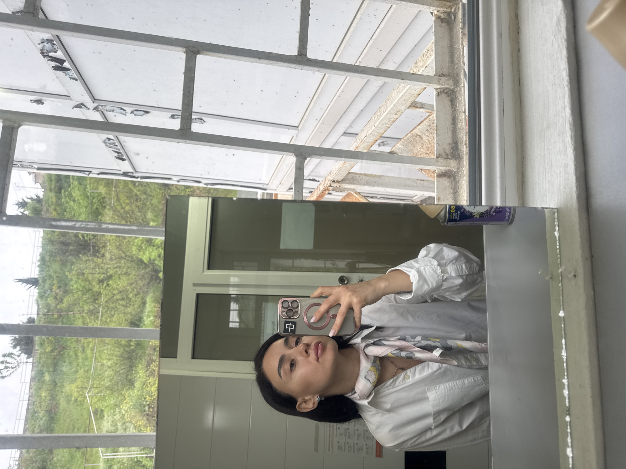 A selfie is being taken in a mirror. The person holding the phone has black hair, is wearing a white collared shirt with a patterned scarf, and the phone case has a Chinese character on it. The reflection in the mirror shows a white tiled wall and a door. Outside the window, a green hillside is visible. The location is not apparent and not evident to be Tbilisi, Georgia.