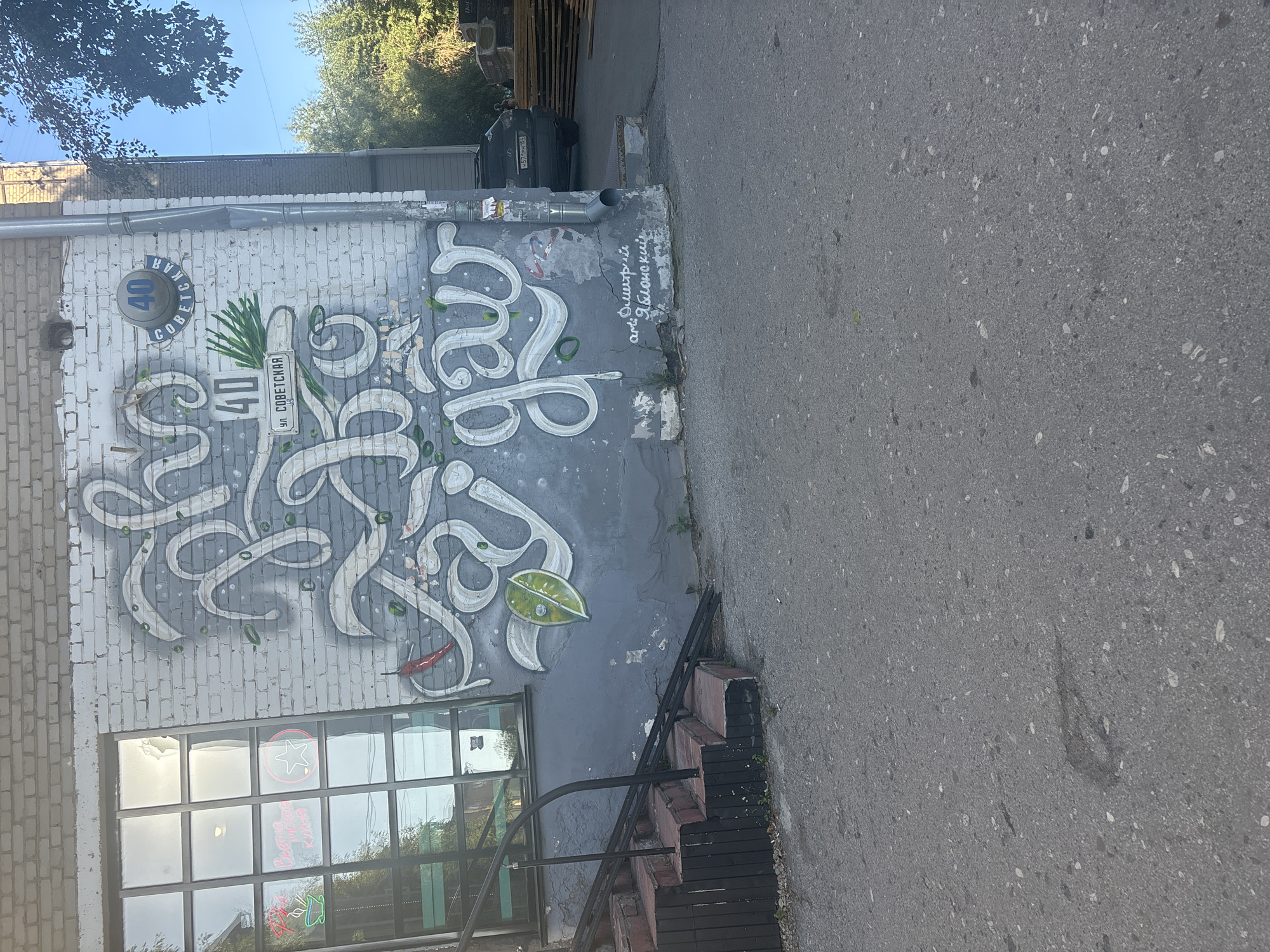 A weathered brick building in Novosibirsk, Russia, features a large, stylized graffiti mural on its exterior wall. The mural, predominantly in white and grey tones, depicts flowing calligraphy with green accents and a red chili pepper. Above the mural, a circular blue sign reads "40" in white, and below it, a rectangular sign with the Cyrillic text "ул. СОВЕТСКАЯ" (Sovetskaya Street). To the left of the mural, a large window with multiple panes reflects the sky and surrounding trees. A metal railing guides visitors up a short set of brick steps to the building's entrance. A dark-colored SUV is parked to the right of the building, with stacks of lumber visible in the background. The ground in front of the building is paved with asphalt. The scene suggests an urban environment with a touch of artistic expression.