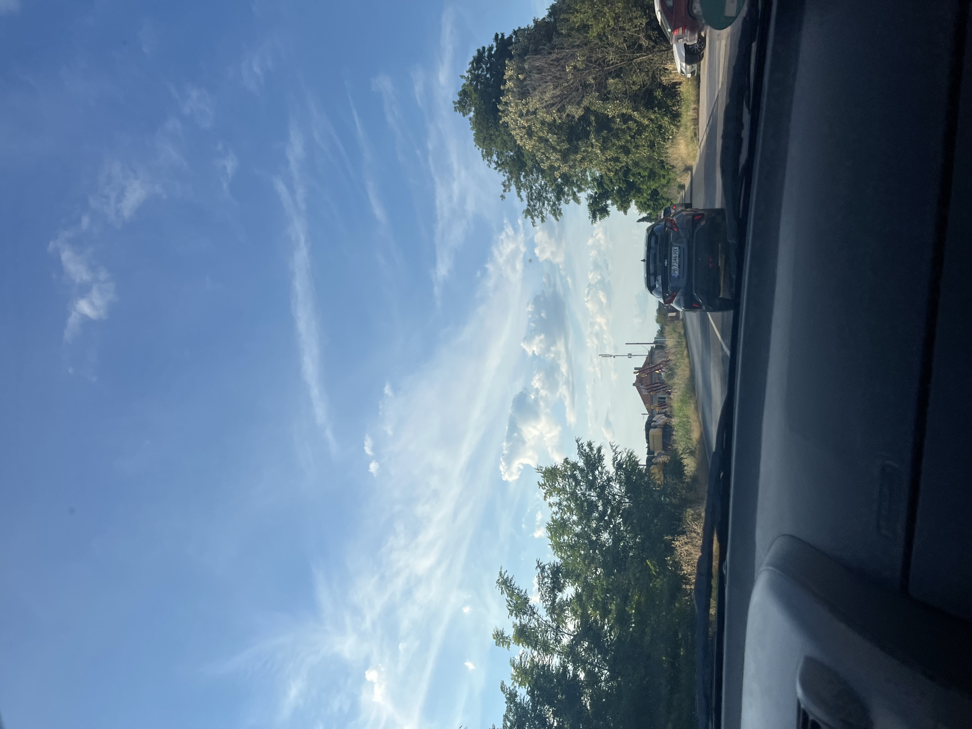 The image shows a view from inside a car, looking forward towards a road in the city of Trud, Bulgaria. The sky is blue with wispy white clouds and streaks of cirrus clouds. On the left side of the road, there are green trees and what appears to be construction equipment, possibly a yellow excavator and red scaffolding on a building. On the right side of the road, there are more trees and parked cars. The car in front of the viewer is a dark-colored hatchback with a license plate visible, "PB 7346 HX". The road surface is asphalt, and there are some dry grass and weeds along the sides of the road. The overall impression is a rural or suburban road scene on a clear day.