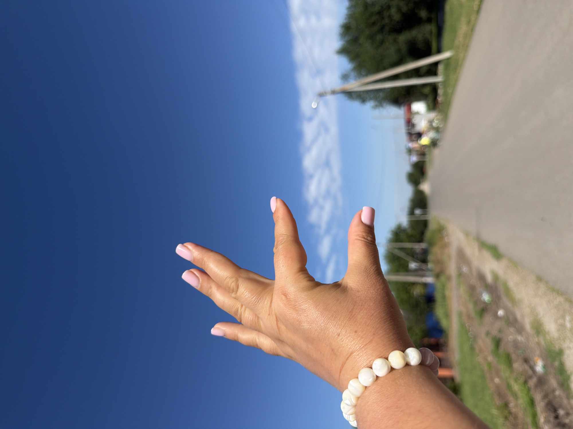 The image is a close-up of a person's left hand, with light-toned skin and light pink fingernails, extended towards the viewer. The hand is adorned with a bracelet made of round, white beads. The background is a bright, clear blue sky with a few wispy white clouds. Below the sky, a paved road recedes into the distance, flanked by green grass and trees. The scene appears to be an outdoor, possibly rural, setting. The location is identified as Leningradskaya, Russia.