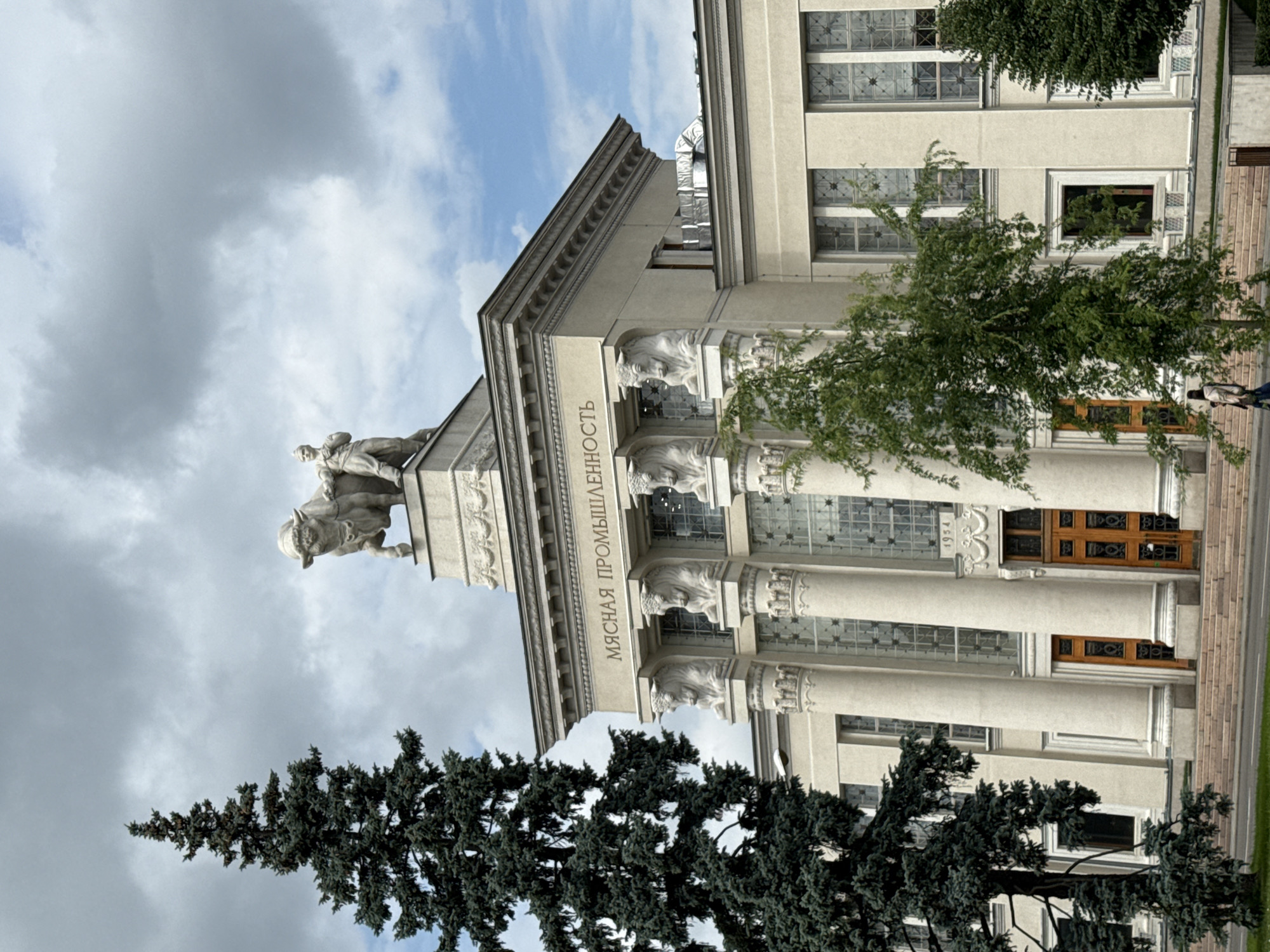 The image captures the Pavilion of the Meat Industry, located at VDNKh in Moscow, Russia. The building features a neoclassical architectural style with grand columns and decorative elements. A prominent sculpture of a man with a bull adorns the rooftop, symbolizing the pavilion's theme. The facade is inscribed with Russian text, "МЯСНАЯ ПРОМЫШЛЕННОСТЬ" (Meat Industry). Two large wooden doors are visible at the base, flanked by staircases. A tall evergreen tree stands to the left of the building, and several deciduous trees are positioned in front, partially obscuring the lower levels. The sky above is filled with dramatic, cloudy formations. A person is walking on the steps in the lower right quadrant of the image. The year "1954" is inscribed above the entrance.