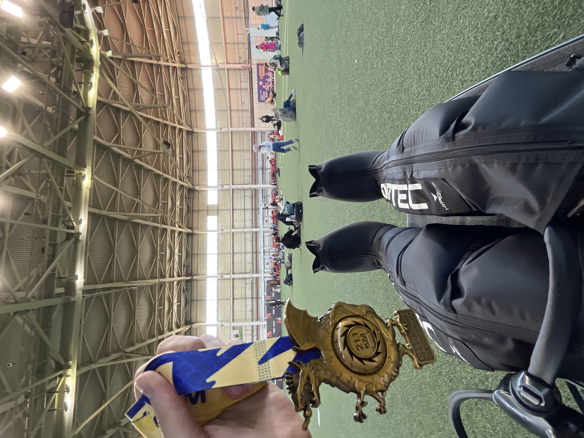 The image depicts an indoor athletic facility with a green turf field and a high, exposed steel beam ceiling. Numerous individuals are scattered across the field. A hand holds a medal on a ribbon, with the inscription "21.1 KM" on the medal, next to black stroller. The stroller brand "Dynatec Hyperlite" is visible on one of the stroller's side pockets. The people, medal, and stroller are in focus. The location is Kar'yer, Russia.