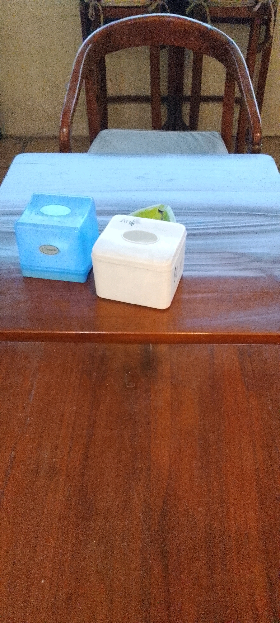 The image shows two tissue boxes on a wooden table. One tissue box is blue and the other is white. A chair with a light-colored cushion is visible in the background. The scene appears to be indoors, likely in a home or restaurant setting. There are no people visible in the image. The location in the city of City of Medan, Indonesia is not evident from the image.
