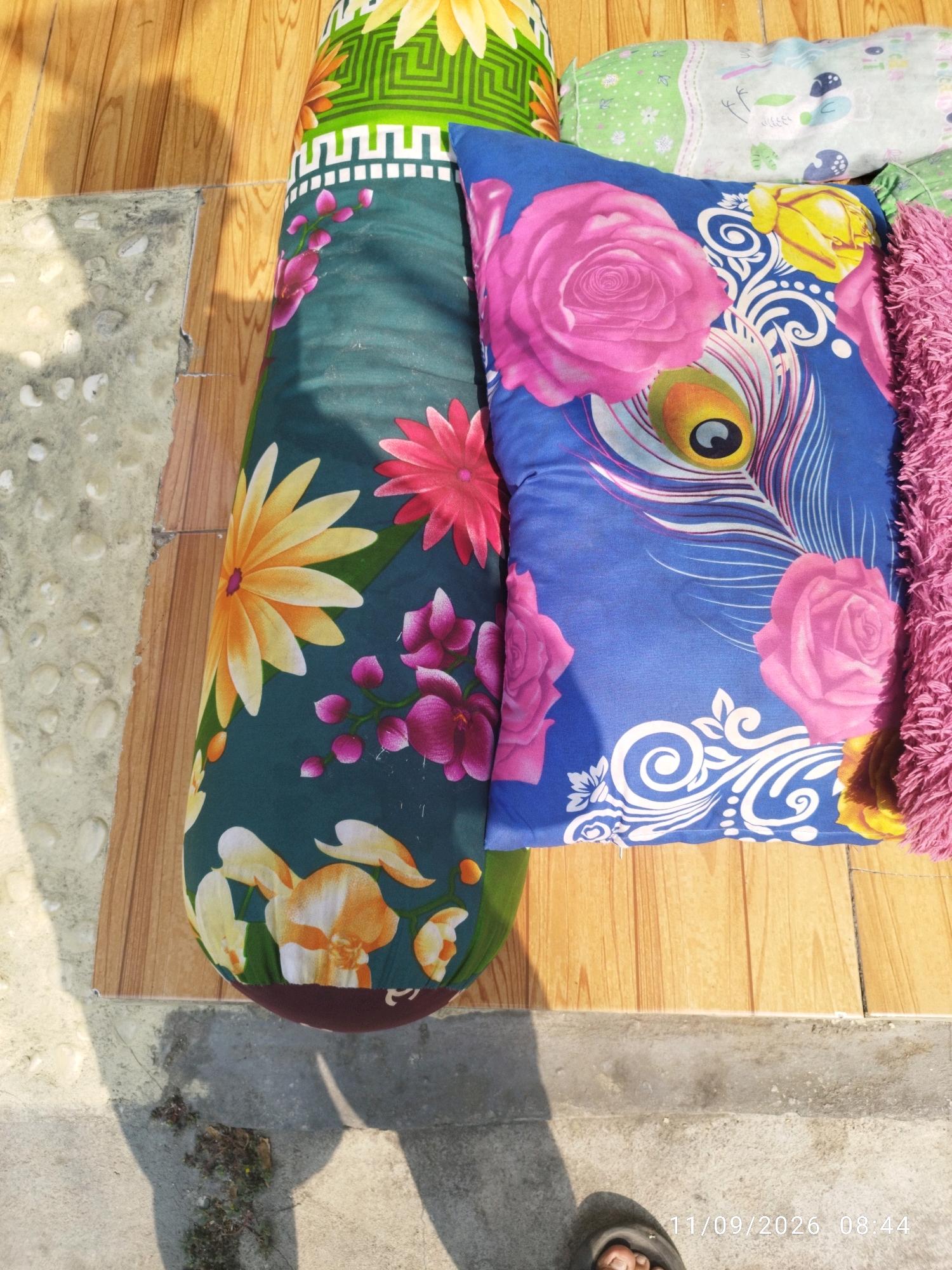 The image shows a pile of colorful pillows and bolsters on a wooden floor, with a concrete border visible on the left. The pillows feature floral patterns, including roses and sunflowers, as well as a peacock feather design on a blue pillow. A pink fuzzy texture is visible on the right side of the image, likely from another pillow or blanket. The lighting suggests it is daytime, with shadows cast on the wooden floor. The lower right corner of the image contains a timestamp reading "11/09/2026 08:44" and a foot wearing a sandal is partially visible, suggesting a personal environment. This scene could be located in a residential area within the city of Mojokerto, Indonesia, possibly an outdoor patio or living space.