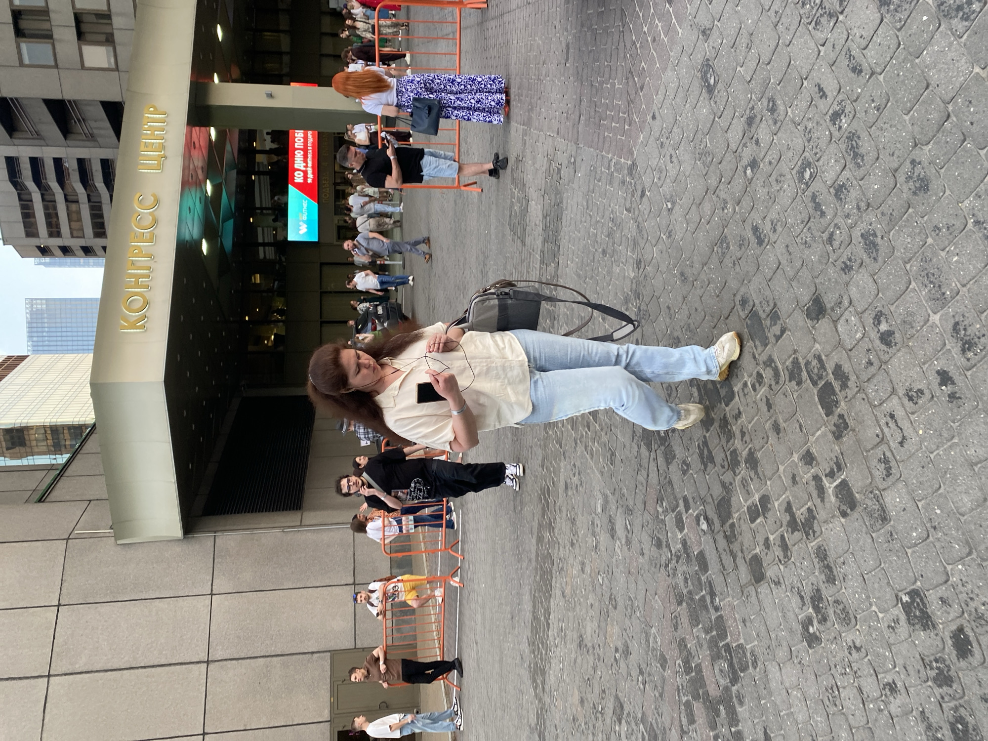 The image shows a scene outside the "Конгресс Центр" in Moscow, Russia. The foreground is dominated by a woman looking at her phone, wearing a light-colored shirt and blue jeans, with a handbag slung over her shoulder. She stands on a paved surface.
Behind her, an orange barrier separates a queue of people, some of whom are looking at their phones as well. There is a mix of ages and clothing styles among the people in the line. A digital display with writing in Russian sits behind the barrier, visible above the heads of some of the queue.
The entrance to the "Конгресс Центр" building is on the left, with its signage clearly visible. A tall building can be seen in the background behind the "Конгресс Центр."