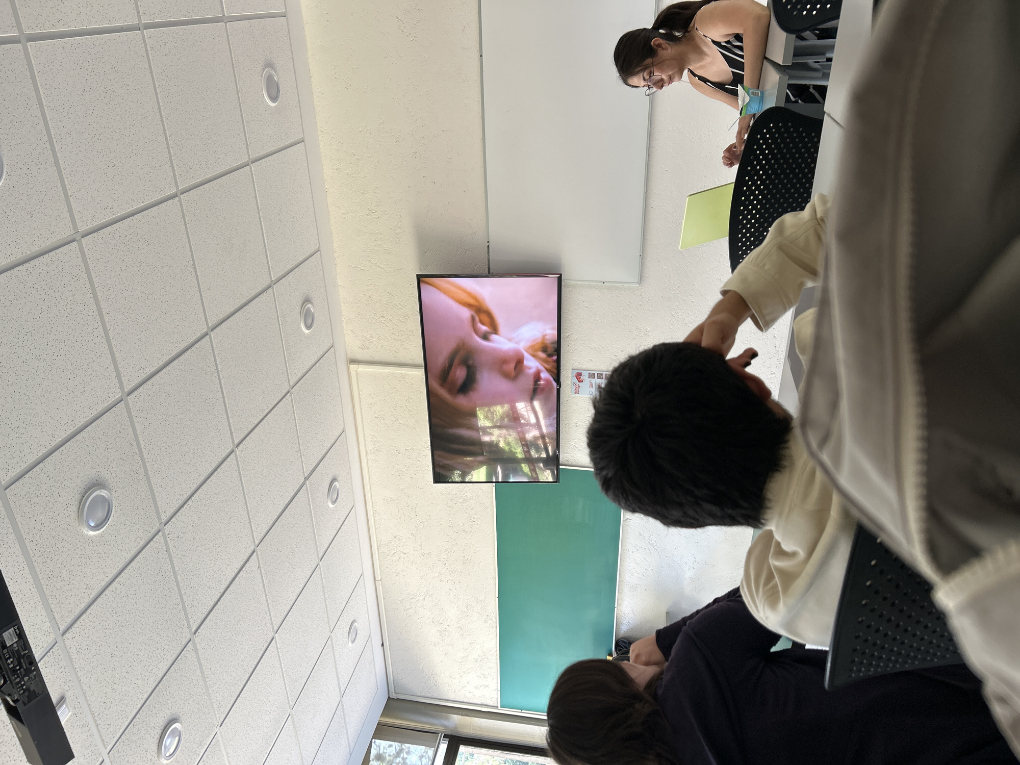 The image appears to be taken in a classroom with a suspended ceiling and recessed lighting. A flat-screen television is mounted on the wall, displaying a close-up image of a blonde woman's face. Below the television is a green chalkboard or whiteboard. Several people are present in the room. A woman in a striped tank top sits near a white board, while other individuals are seated at desks, partially visible. The perspective is from a low angle, suggesting the photographer is seated or positioned lower than the subjects. The location, San Bernardino Tlaxcalancingo, Mexico, cannot be confirmed from the visual information.
