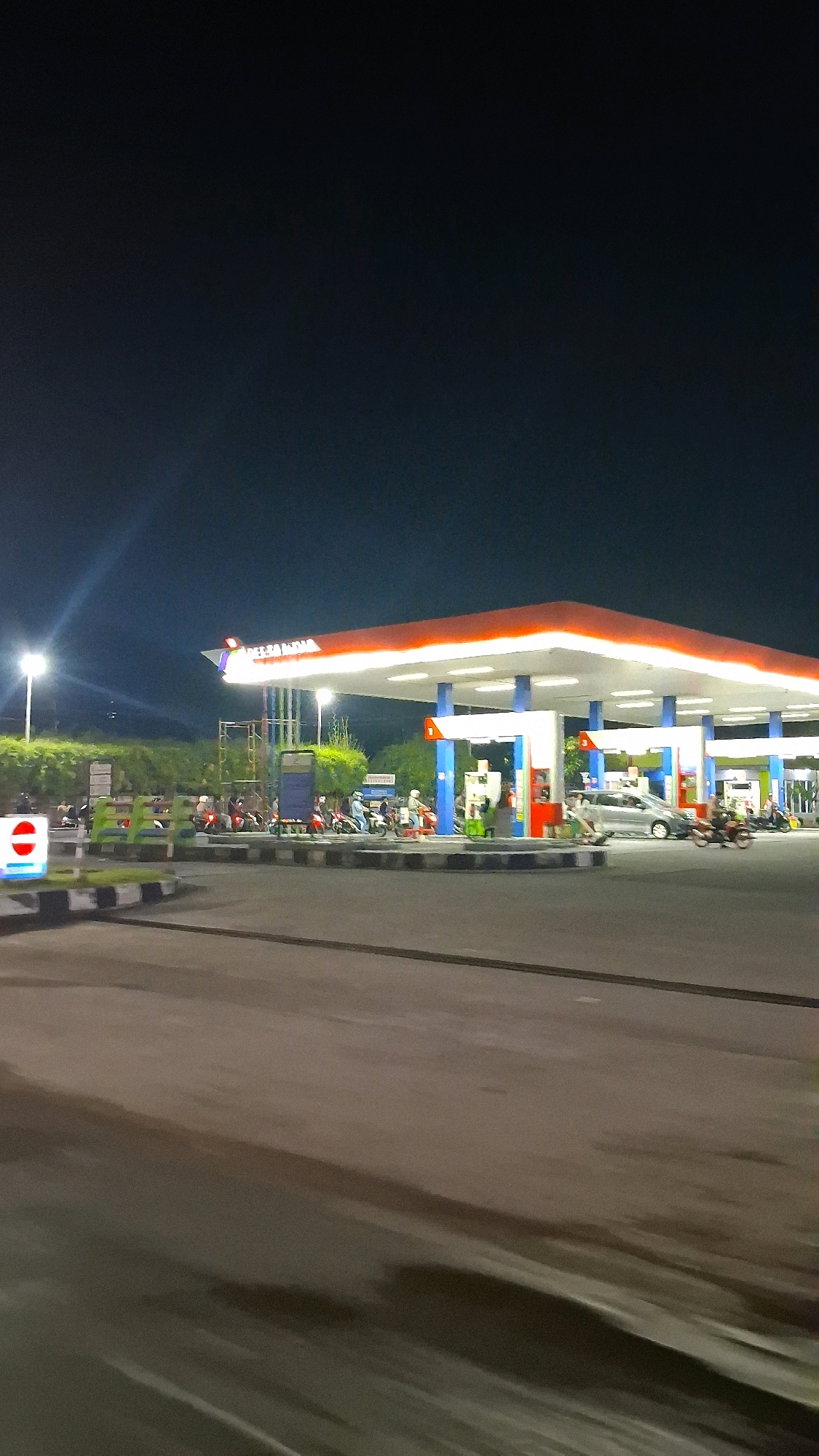 The image depicts a nighttime scene at a Pertamina gas station in Berbah, Indonesia. The gas station is illuminated by bright overhead lights, casting a glow on the surrounding area. Multiple vehicles, including cars and motorcycles, are present at the pumps, suggesting ongoing activity. Several people are visible, either attending to their vehicles or standing near the pumps. The gas station features a prominent red and white canopy, supported by blue pillars, a common design for Pertamina stations. In the foreground, a portion of the road is visible, with white and black striped markings indicating a pedestrian crossing or traffic lane divider. The background is dark, consistent with a night setting, with some trees and structures barely discernible. A sign with a red circle and a horizontal bar, often signifying "no entry" or "stop," is visible to the left of the gas station. The overall impression is one of a busy, functional gas station operating during the night.