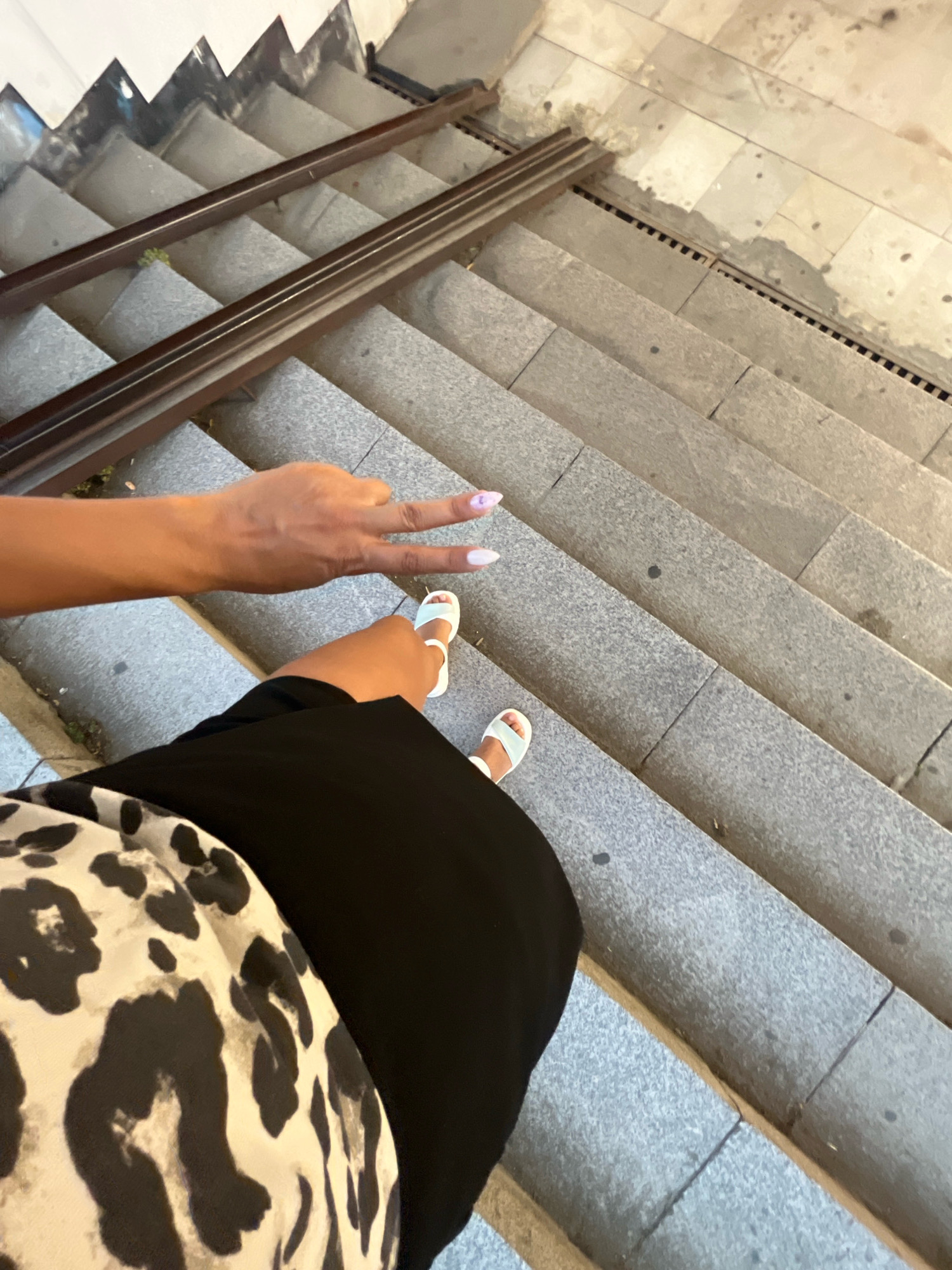 The image shows a first-person perspective of someone standing on outdoor granite stairs in Plovdiv, Bulgaria. The person is wearing white sandals and a black skirt, and a top with a black and white animal print pattern. Their left hand is raised, forming a peace sign. There are dark brown metal handrails on the stairs, and the stairs themselves appear slightly worn. The environment suggests an urban setting, possibly a public space or pathway.