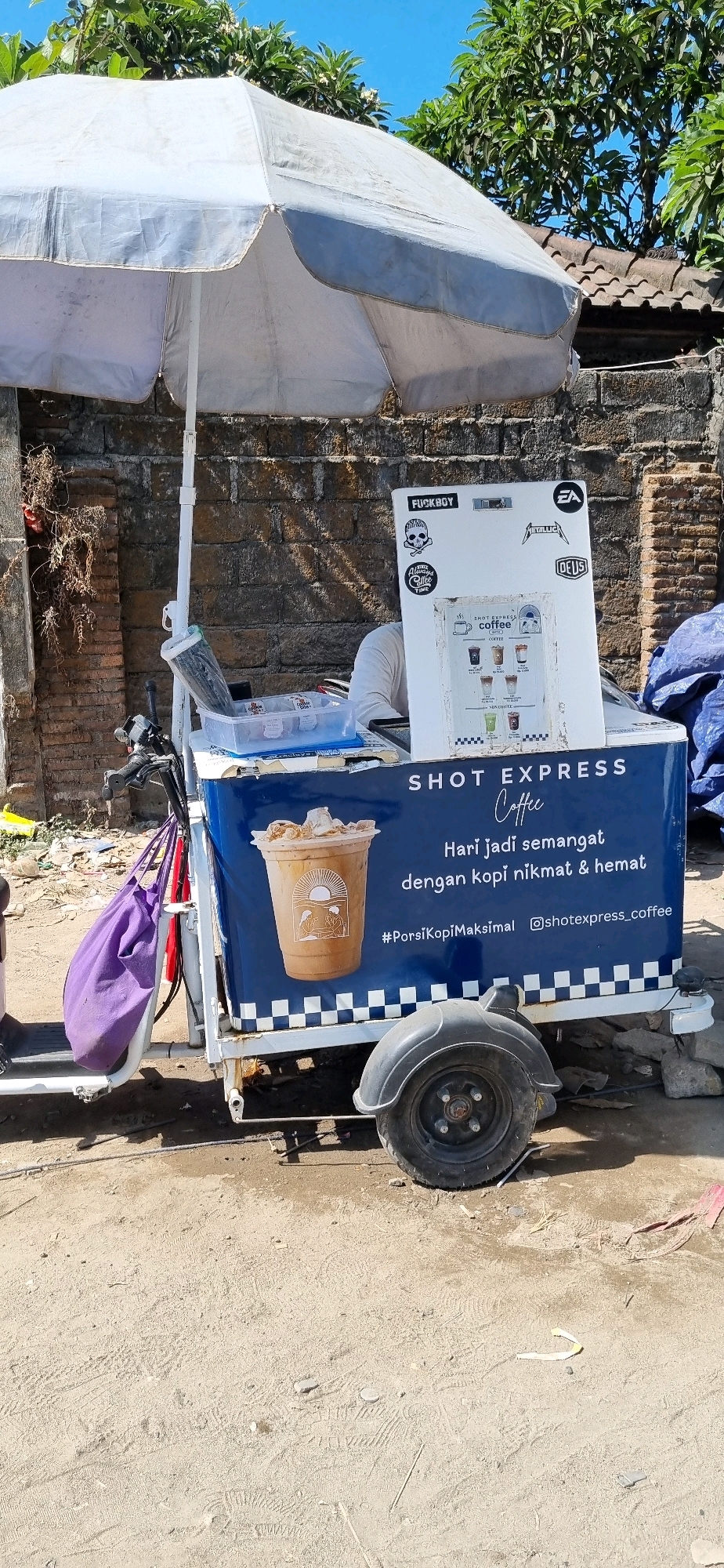 The image shows a mobile coffee cart, likely located in Denpasar, Indonesia. The cart is white and blue, with a large umbrella providing shade. A menu board displays different coffee options and a picture of a coffee cup with a sun and two figures. The cart is positioned on a dirt path, with a brick wall and lush green trees in the background. A purple bag hangs from the handlebars of the cart. The overall scene suggests a street-side vendor operation in a warm, sunny climate.
