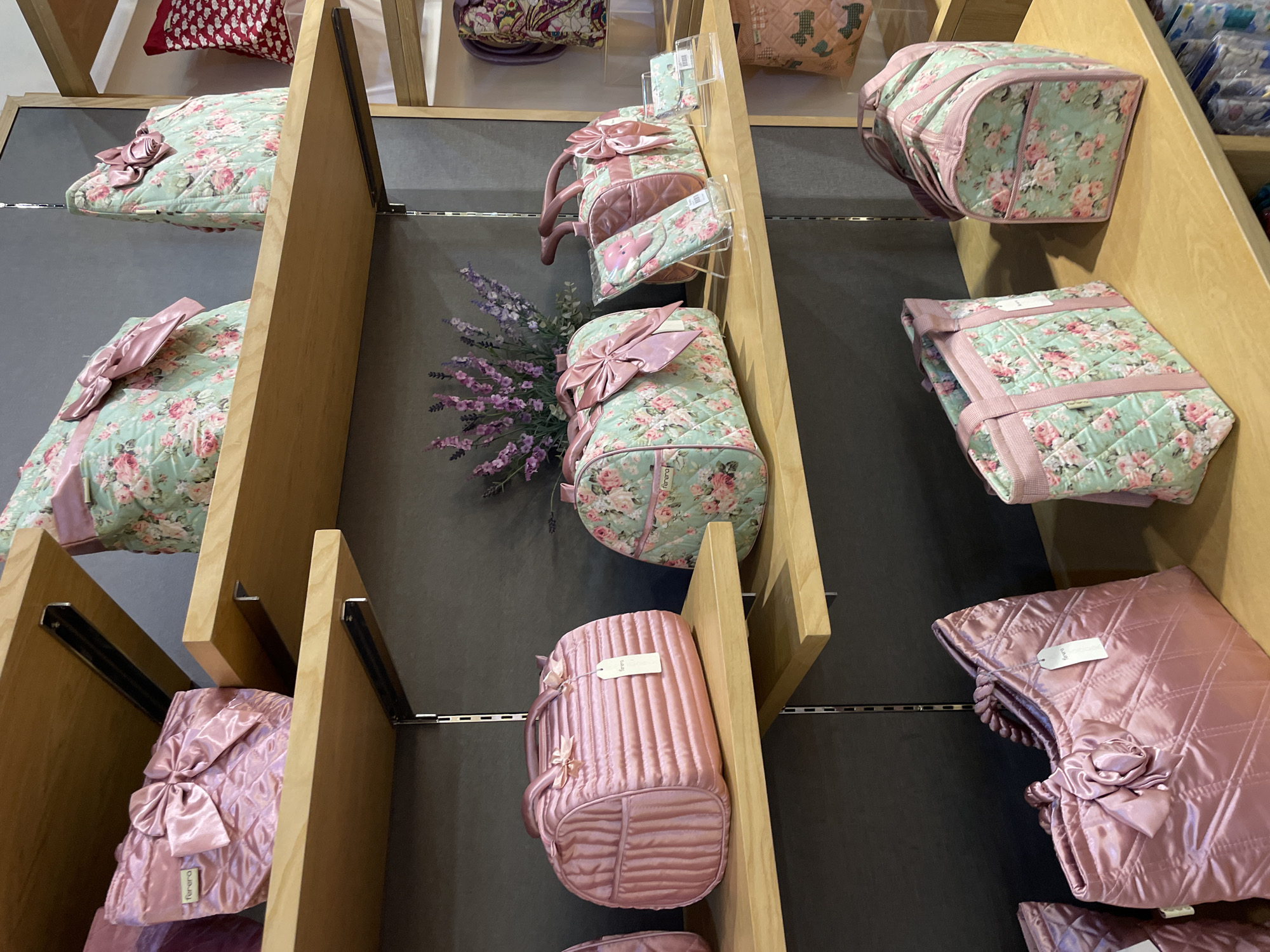 This is a medium shot, eye-level, interior photograph of a retail display of quilted bags on wooden shelves. The bags are in various sizes and colors, with prominent floral patterns and pink bows. The display appears to be in a boutique or department store. The background is a dark gray wall. The lighting is bright and even. The scene is static, with no people or activity.