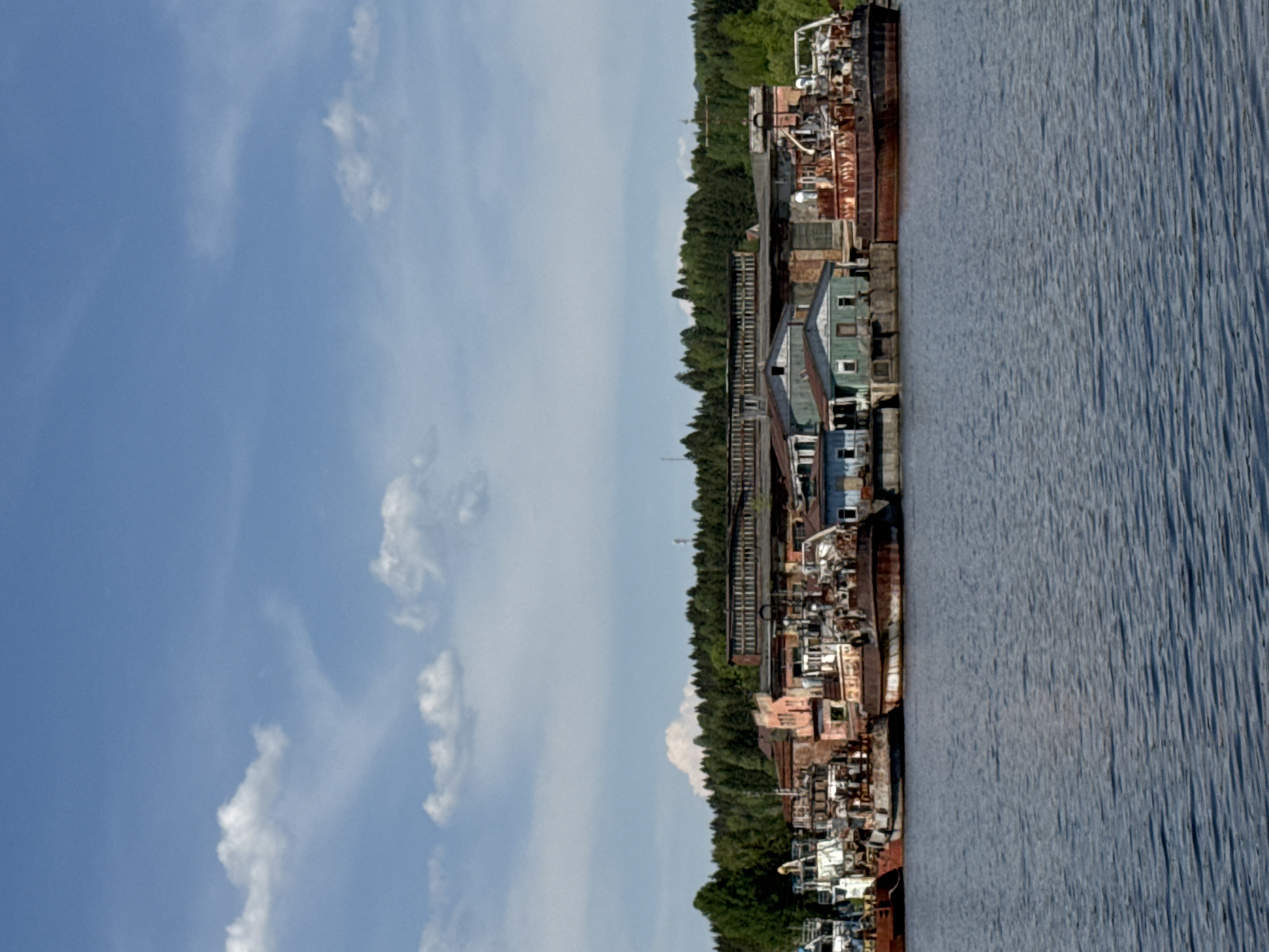 The image depicts a riverside scene in Perm, Russia. A body of water is on the right, displaying prominent ripples. Adjacent to the water is a collection of industrial structures, including buildings and what appears to be a railway line cutting through trees. The scene extends into a dense tree line, leading into a bright, partially cloudy sky on the left. The buildings and structures appear old and weathered, suggesting an aging industrial area. No people are visible in the image.