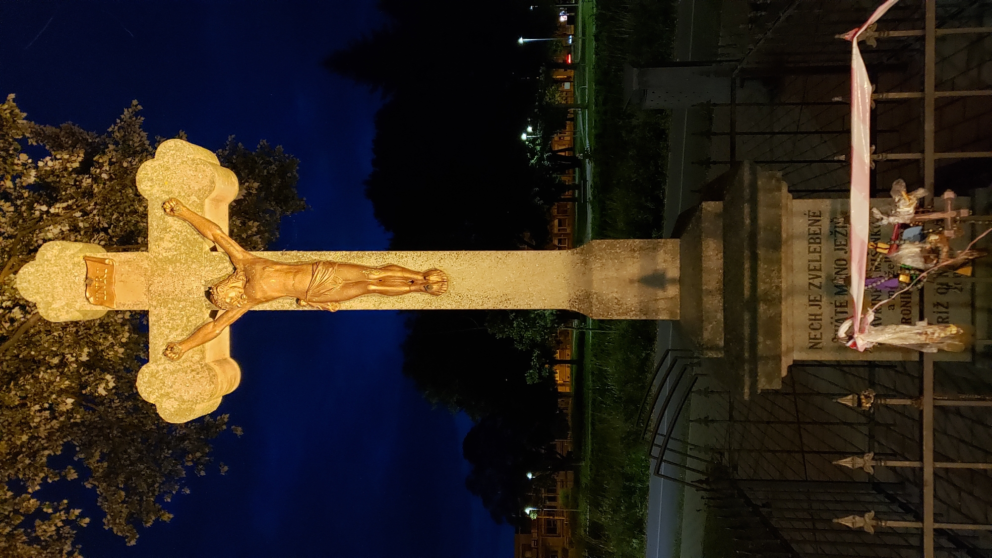 A gilded statue of Jesus Christ on the cross is illuminated against a dark blue sky. The cross is made of weathered stone and is mounted on a substantial stone pedestal. Inscriptions are visible on the base of the pedestal, but they are partially obscured by offerings of ribbons and small trinkets. The cross is located in a grassy area, possibly a park or a roadside verge, with trees and streetlights visible in the background. A metal fence encloses the immediate area around the cross. The scene appears to be in the city of Prešov, Slovakia, indicated by the context provided.