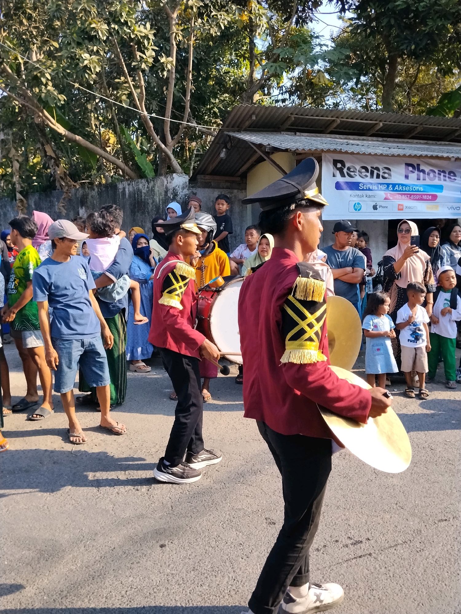 In Tlogosari, Indonesia, a street scene depicts a marching band with children and adults watching. Two young boys in maroon marching band uniforms are in the foreground, one holding a cymbal and the other a drum. They are on a paved road, casting shadows. Behind them, a crowd of spectators, including adults and children, are gathered. A banner for "Reena Phone Servis HP & Aksesoris" is visible in the background, indicating a commercial establishment. The environment is outdoors, with trees and a building with a corrugated roof visible. The overall atmosphere suggests a community event or parade.