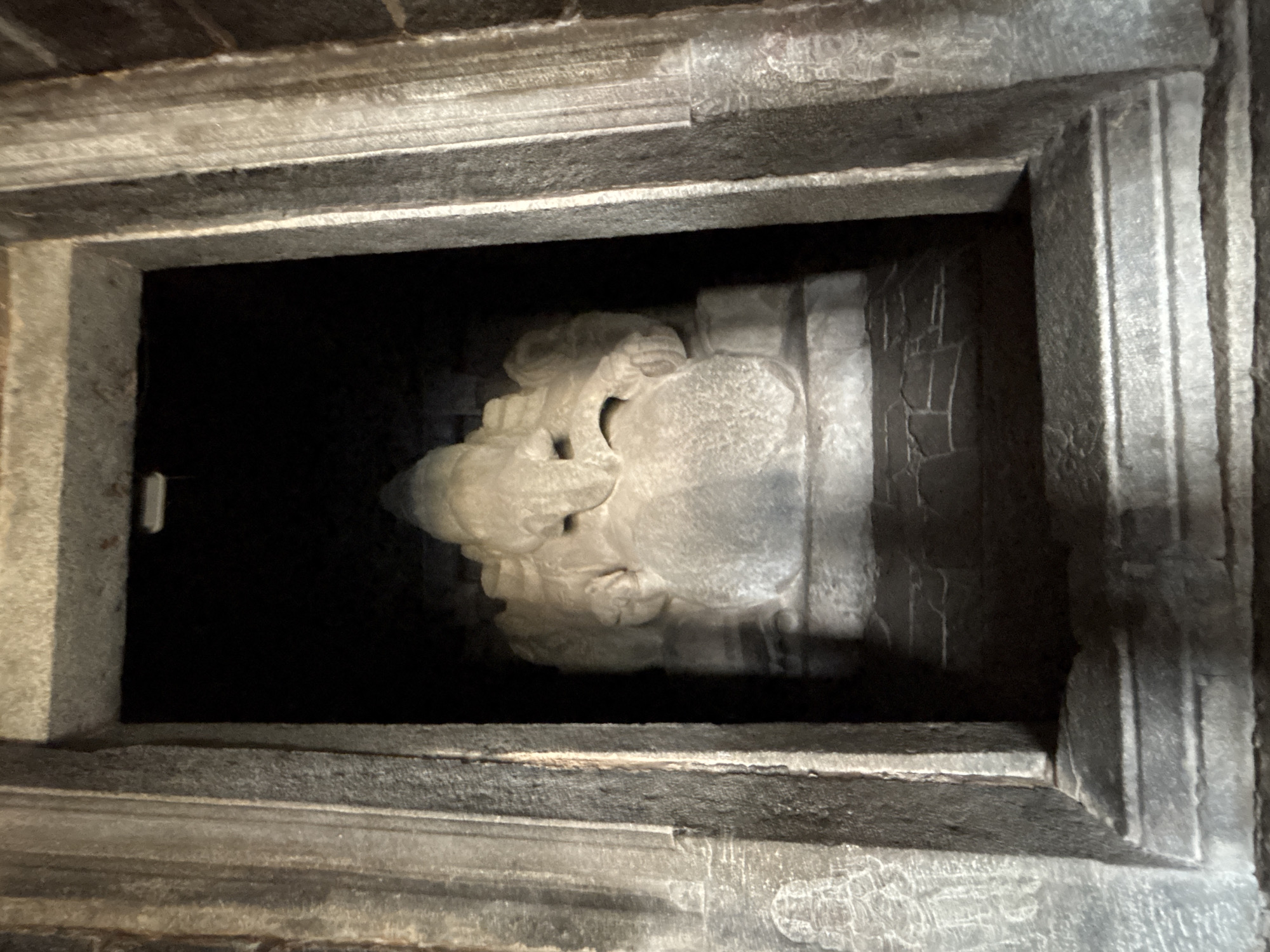 A close-up shot inside a dark, stone-carved temple structure, possibly in Hampi, India, reveals a carved sculpture within a recessed alcove. The alcove is formed by gray stone with detailed carvings on the front facing surfaces. The sculpture, also carved from the same stone, appears to depict a deity or mythological figure with intricate details. The lighting is dim, accentuating the texture of the stone. The scene is static and suggests an ancient and sacred space.